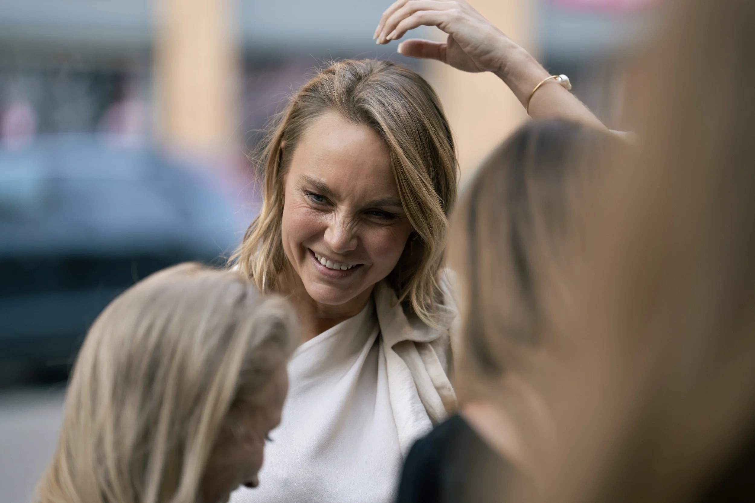 A woman smiling and raising her hand, engaging with three other women who are partially visible in a social outdoor setting.
