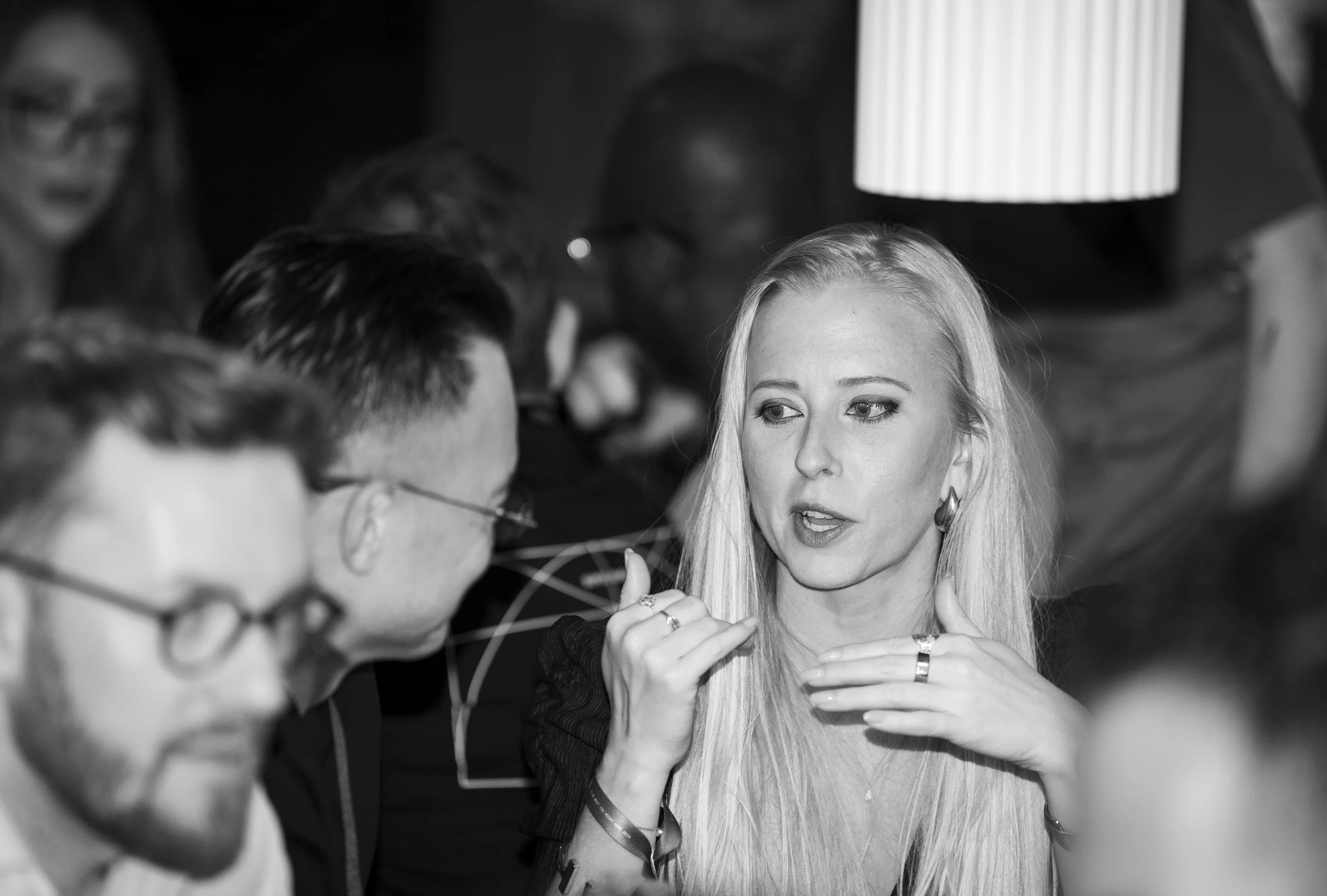 A group of people engaged in conversation, with a blonde woman in focus speaking and gesturing with her hand, in a social setting.