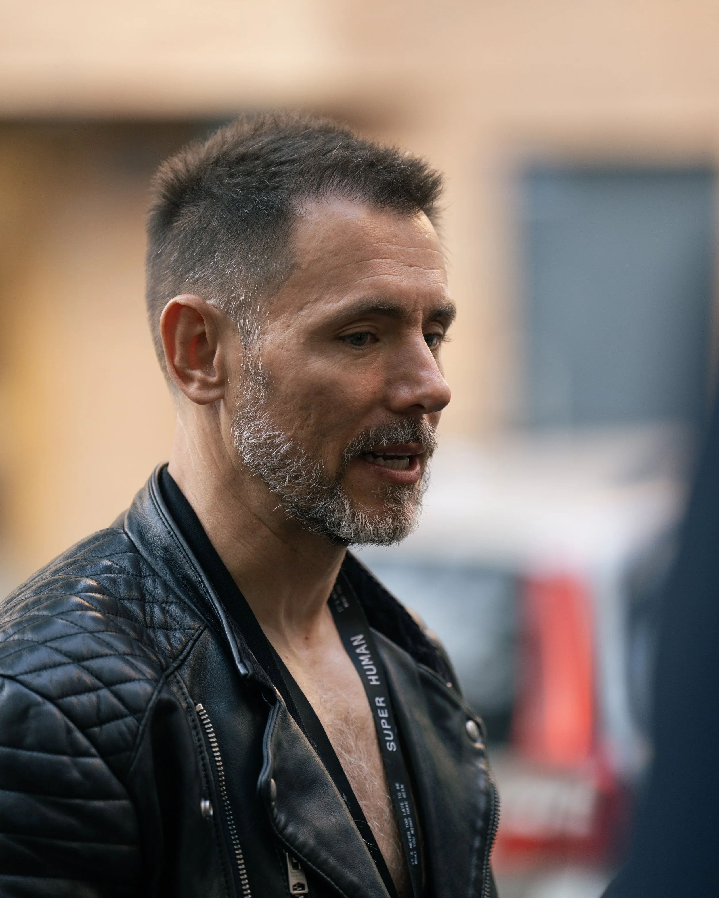A man with short dark hair and a gray beard, wearing a black leather jacket and a lanyard, is speaking or engaged in conversation indoors.