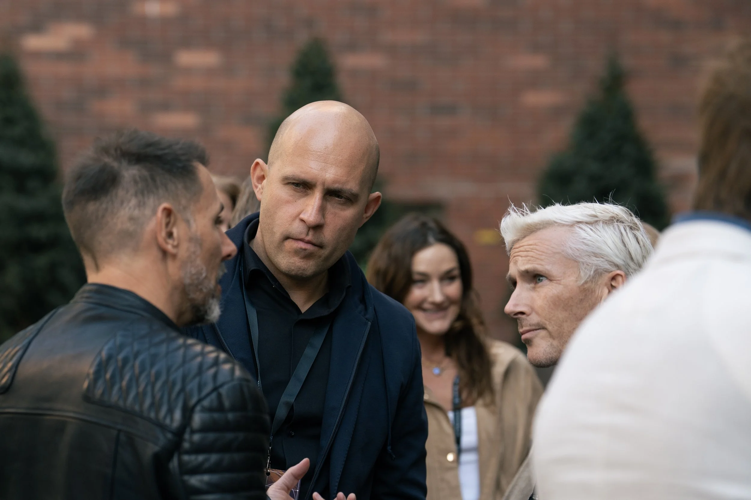 A group of people, including three men and two women, are engaged in a serious conversation outdoors with a brick wall and trees in the background.