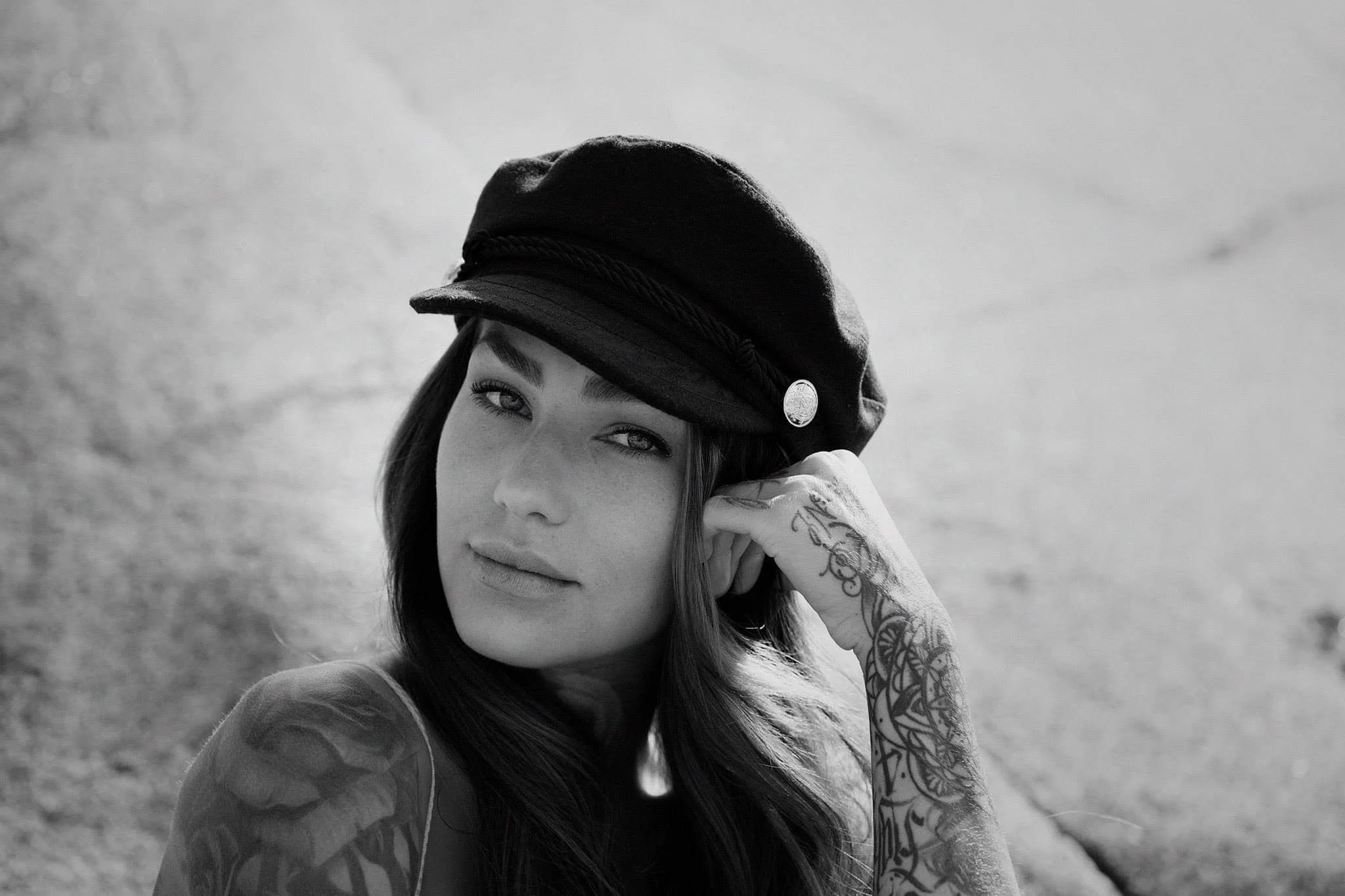 A black-and-white photo of a young woman with tattoos on her arm, wearing a dark hat with a decorative button, touching her ear, with a textured wall in the background.