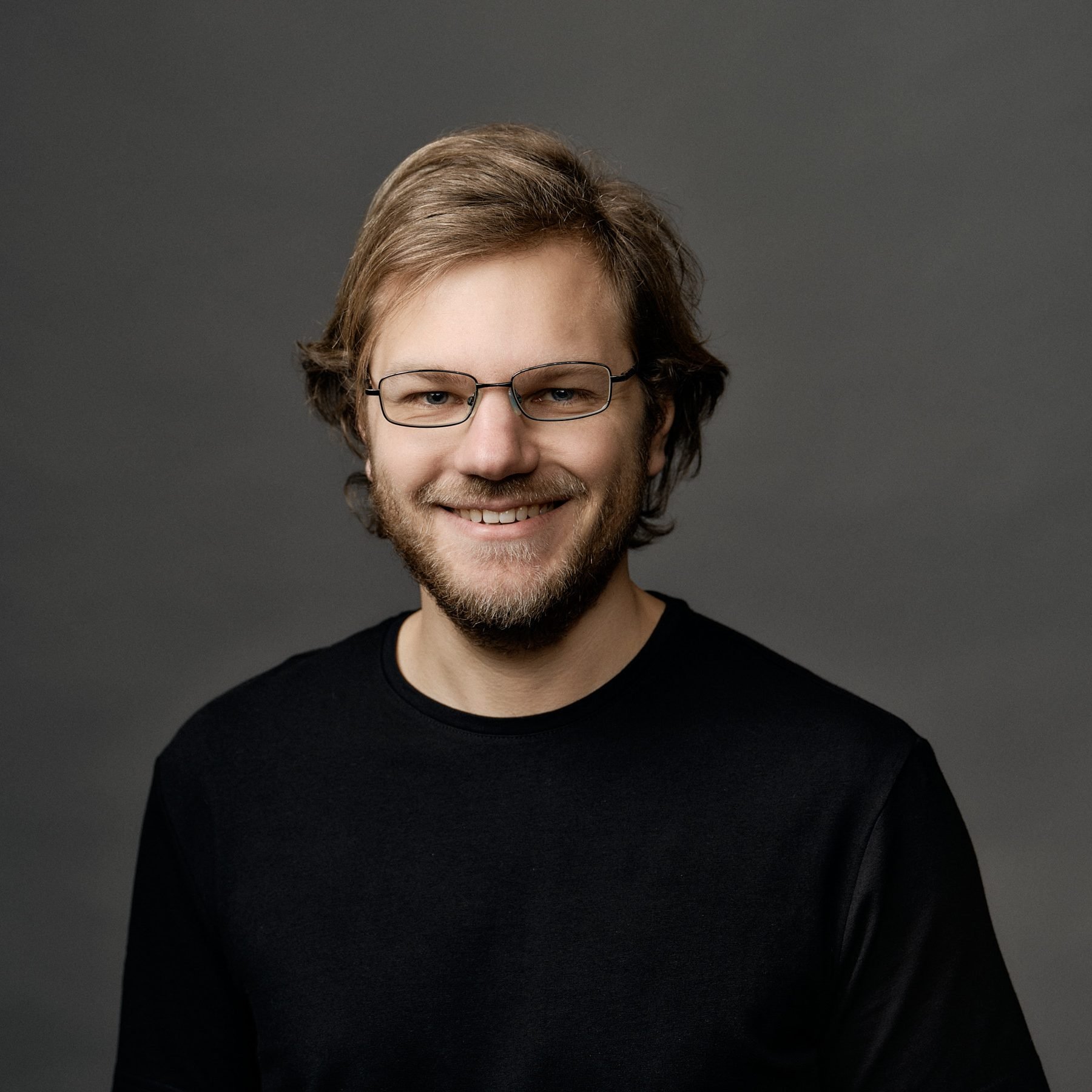 Man with glasses, beard, and long hair smiling, wearing a black shirt against a gray background.