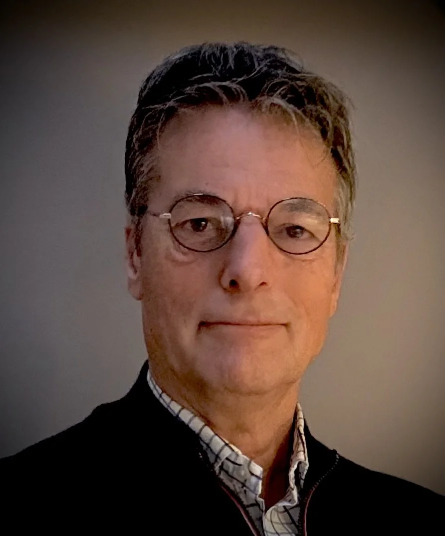 A portrait of a middle-aged man with gray hair wearing glasses, a checkered shirt, and a dark sweater, against a plain gray background.