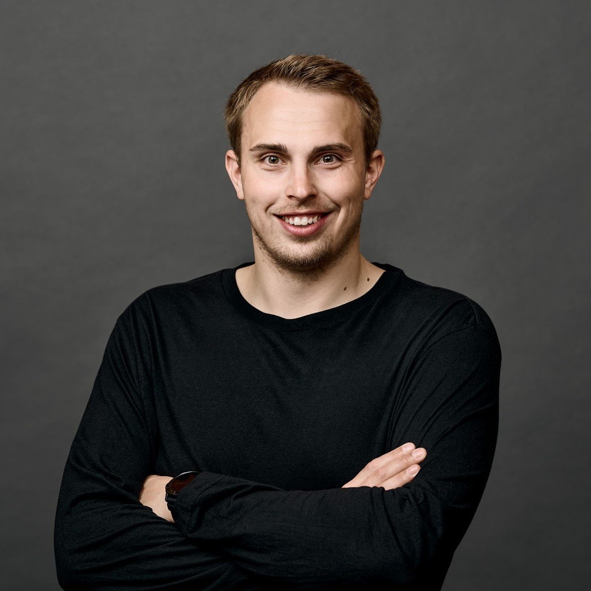 Smiling man in black shirt with crossed arms against gray background.