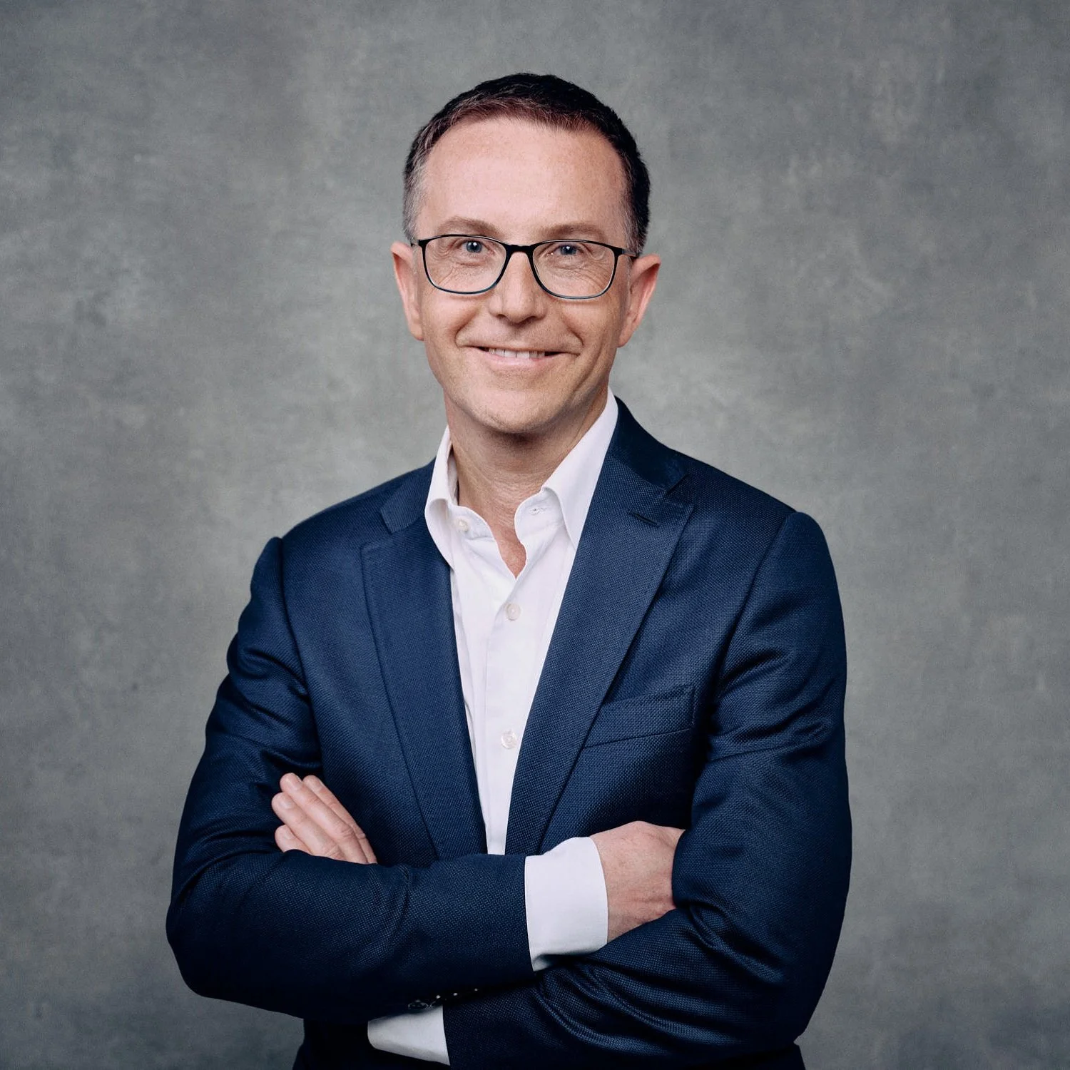 Man in a suit with glasses and arms crossed, neutral background.