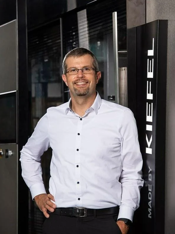 Man in glasses and white shirt smiling next to Kiefel machine.