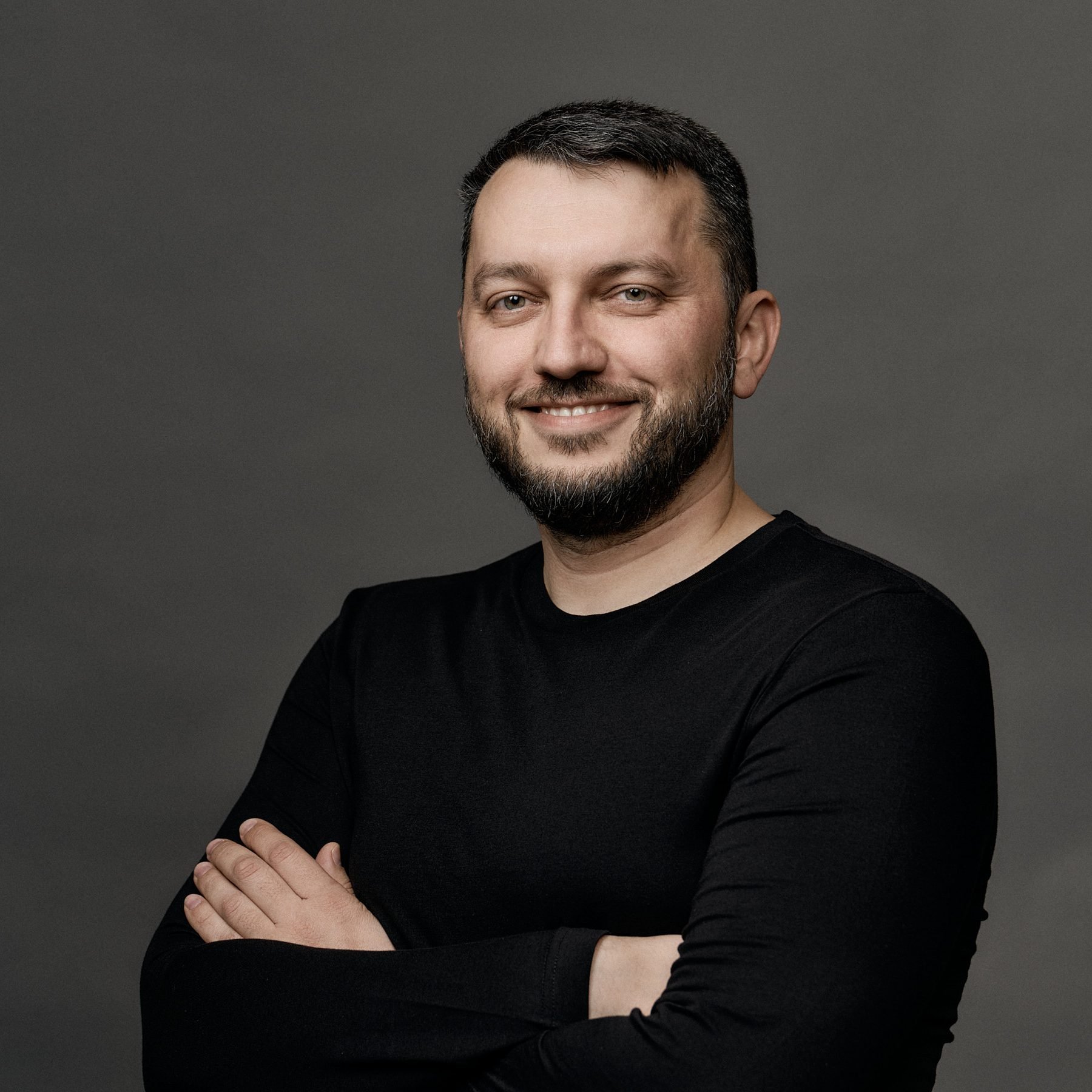 Man with a beard in a black long-sleeve shirt smiling, arms crossed.