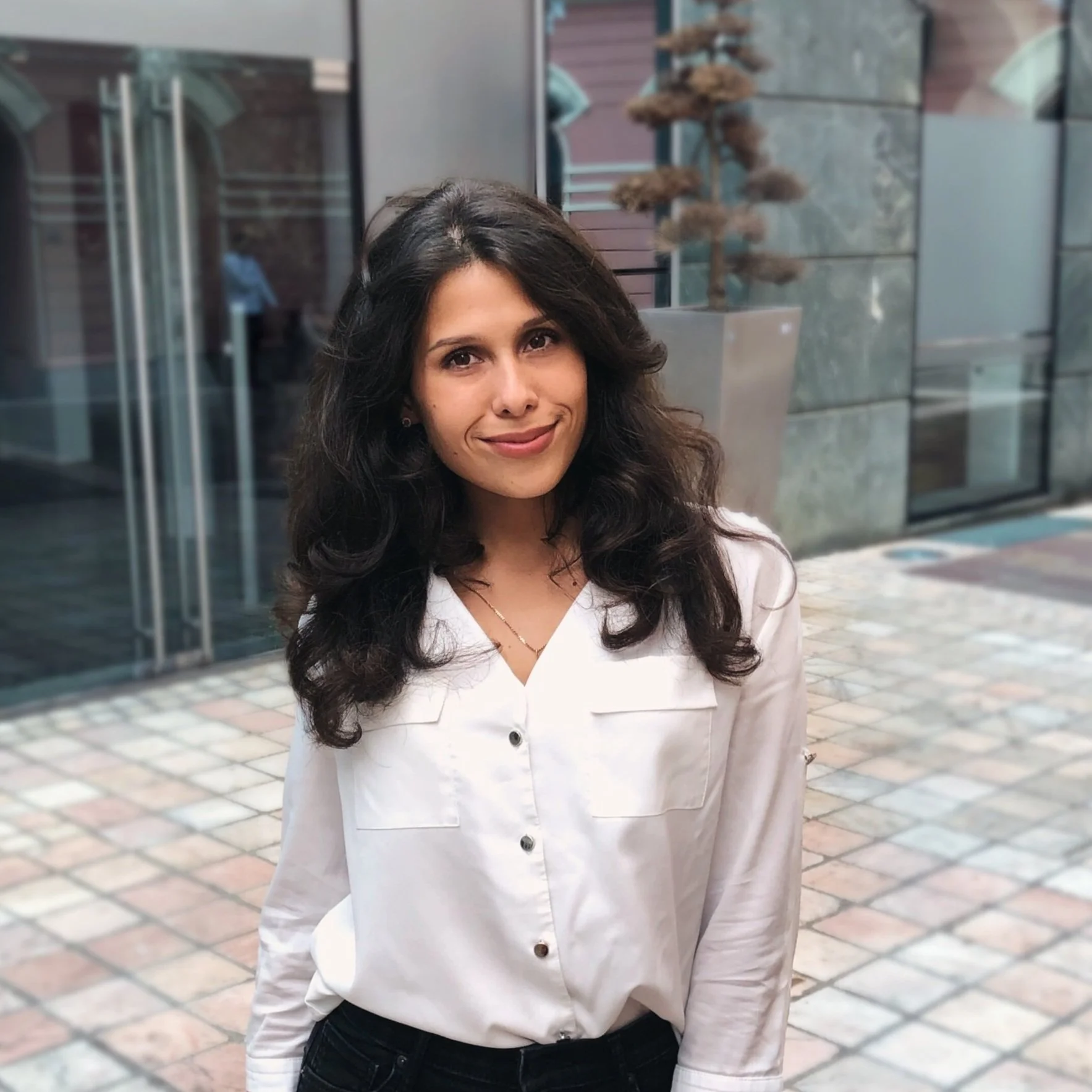 Person wearing a white blouse standing in an outdoor paved area with glass doors in the background.