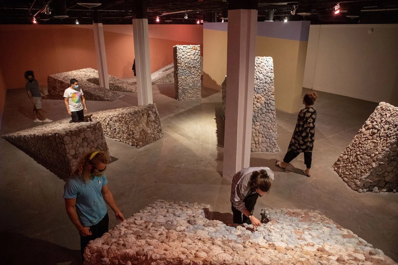Contemporary art installation titled 'Future Pacific' featuring monumental sunken arches covered in clay forms like seashells and corals, displayed inside a gallery with dark ceiling, spotlights, and painted walls in orange, blue, and white.