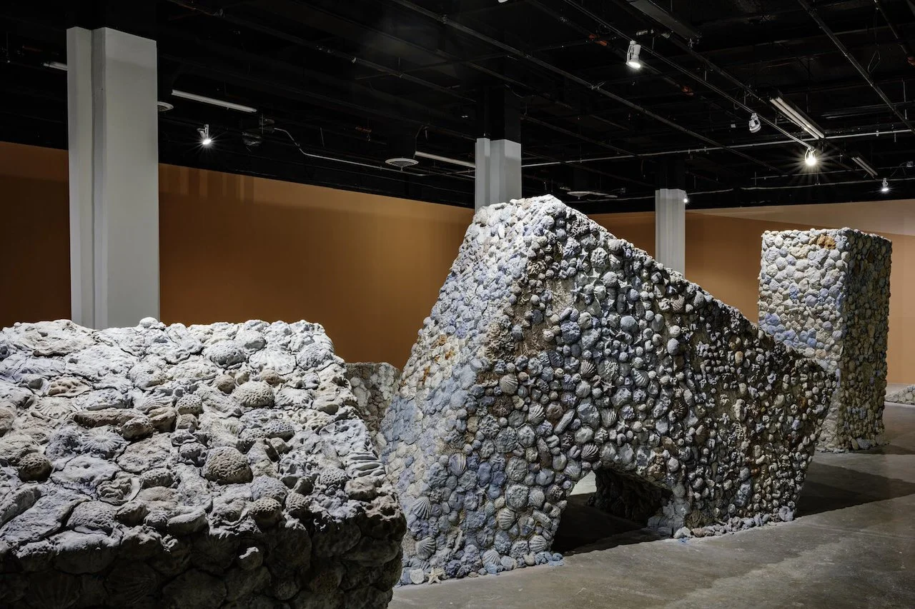 Contemporary art installation titled 'Future Pacific' featuring monumental sunken arches covered in clay forms like seashells and corals, displayed inside a gallery with dark ceiling, spotlights, and painted walls in orange, blue, and white.