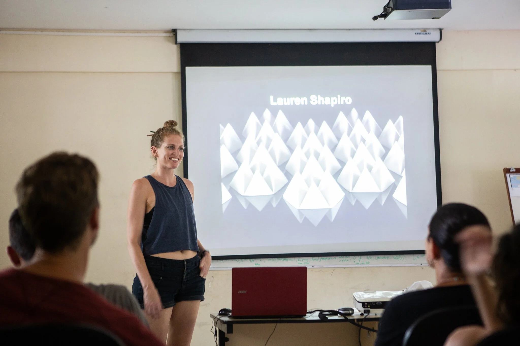 A woman in a sleeveless top and shorts giving a presentation in a classroom, with a slide showing her name, Lauren Shapiro, projected behind her. Several students are seated facing her.