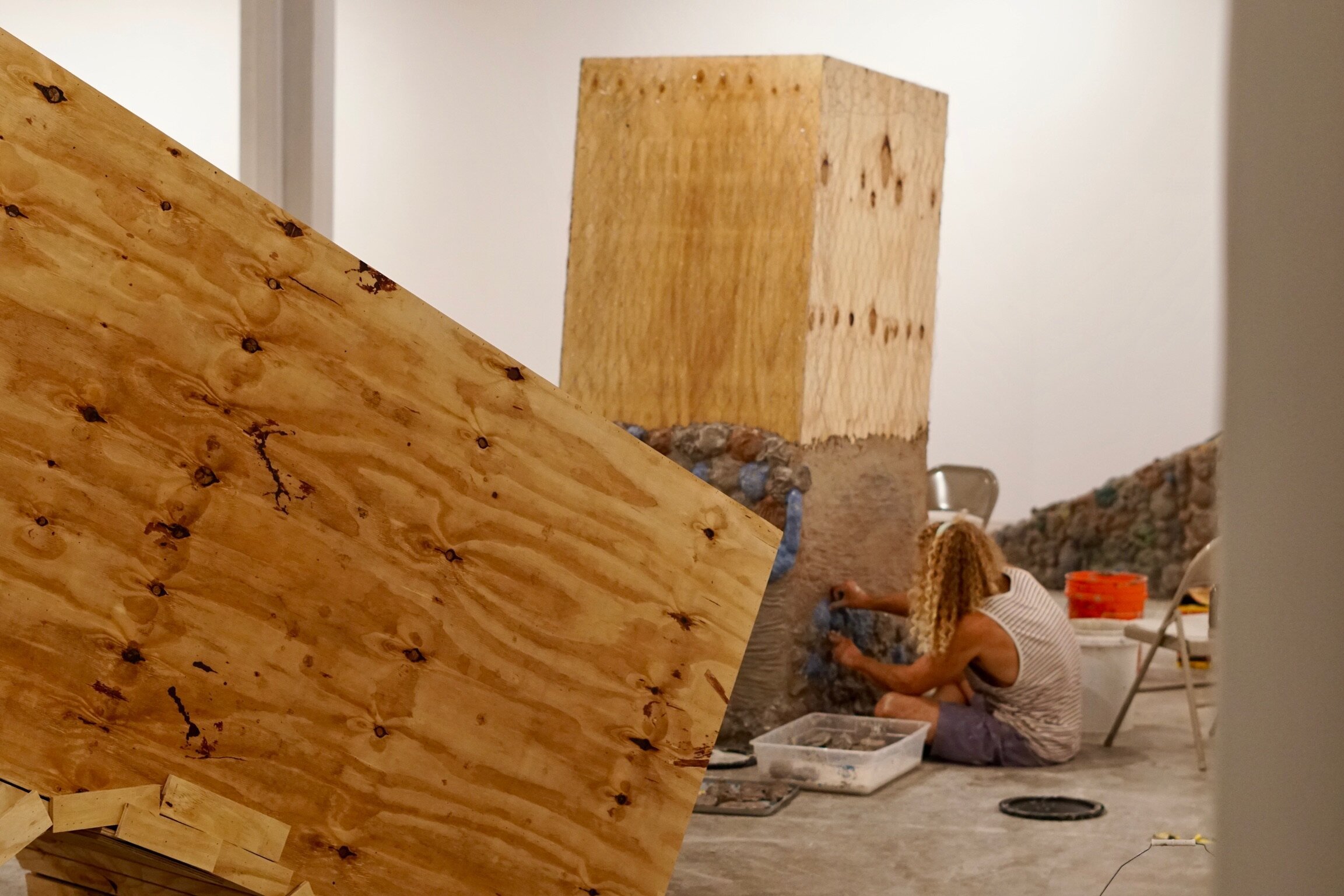 Person working on a large wooden sculpture or installation in anindoor art gallery, sitting on the floor with tools and materials around.