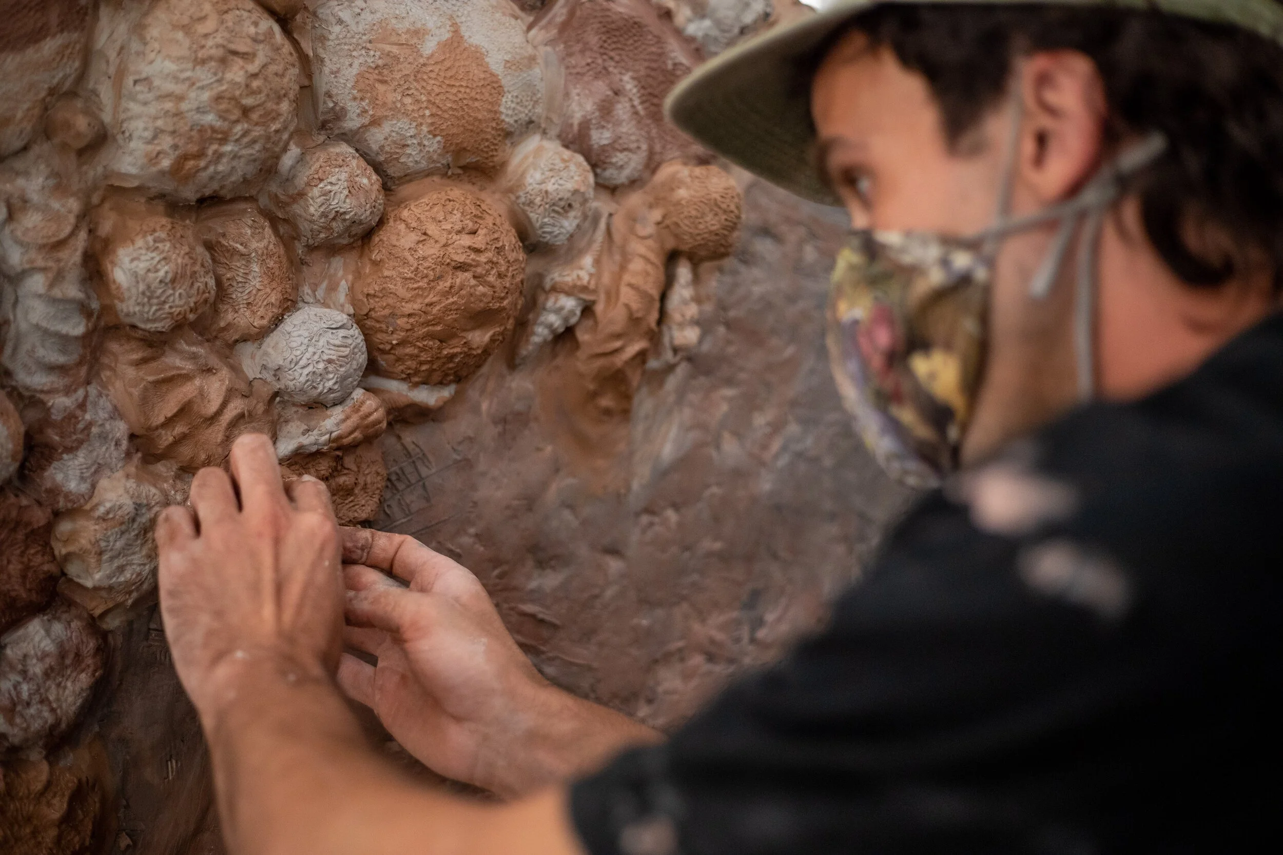A person applying clay seashells, corals, and marine forms to a wall, wearing a hat and a face mask.
