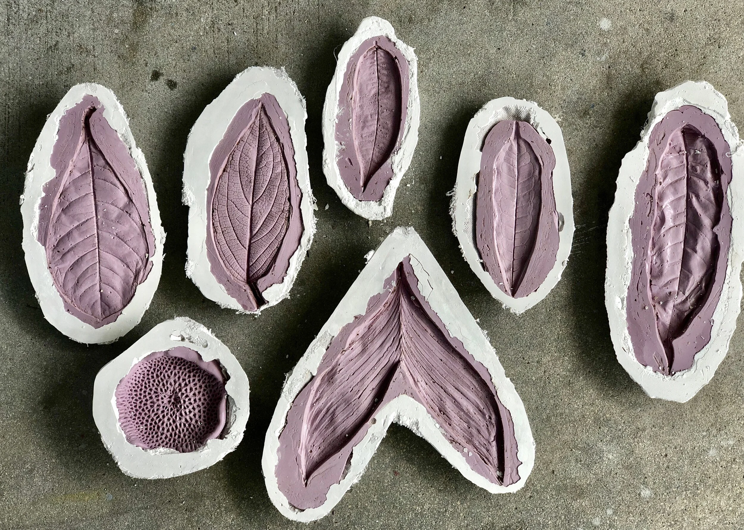 Set of seven decorative clay tiles with carved leaf patterns in purple tones, arranged on a concrete surface.