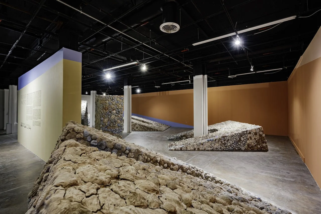 Contemporary art installation titled 'Future Pacific' featuring monumental sunken arches covered in clay forms like seashells and corals, displayed inside a gallery with dark ceiling, spotlights, and painted walls in orange, blue, and white.