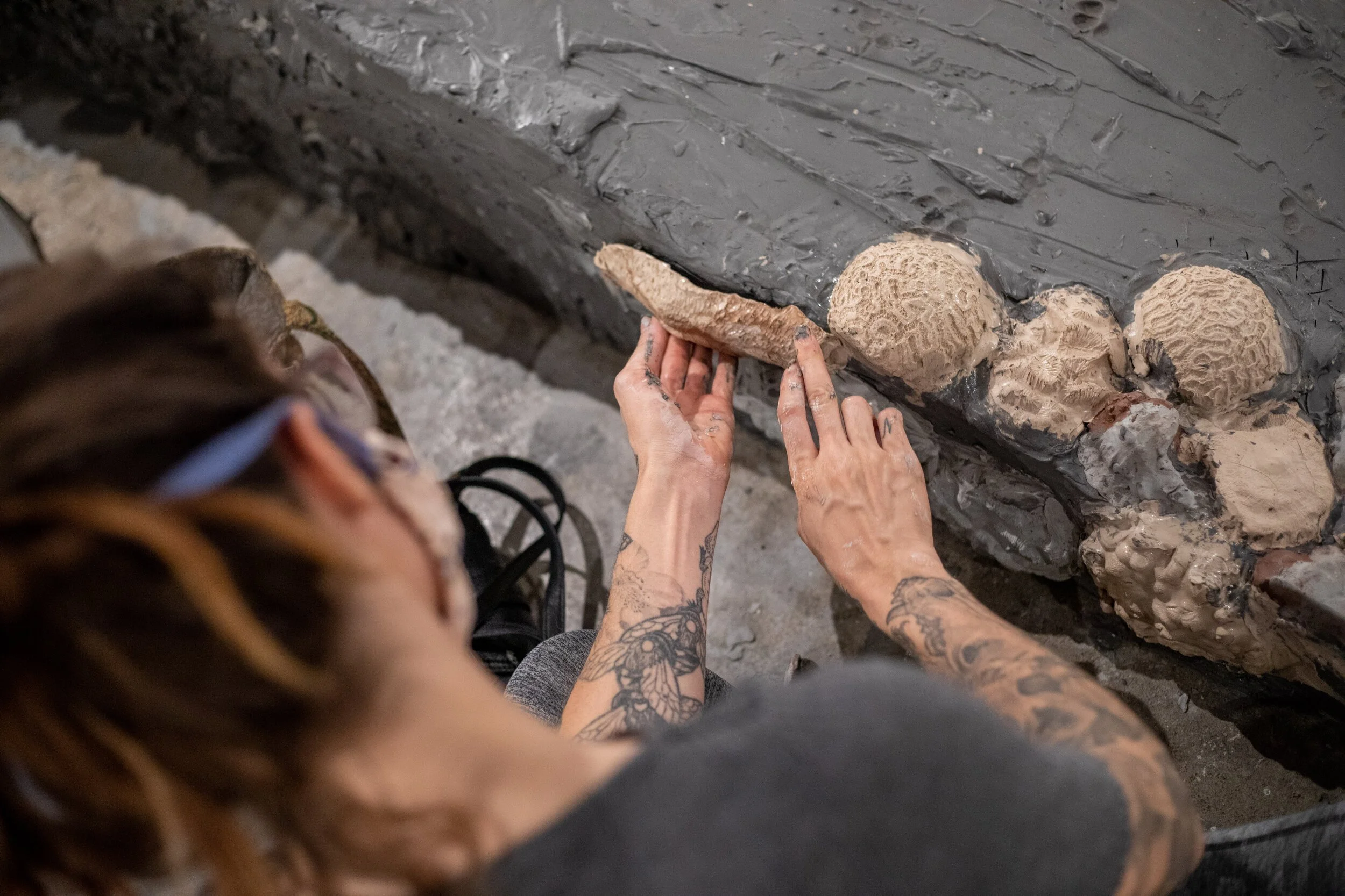 A person with tattoos on their arms is installing shell-like appliques onto a stone or concrete surface.