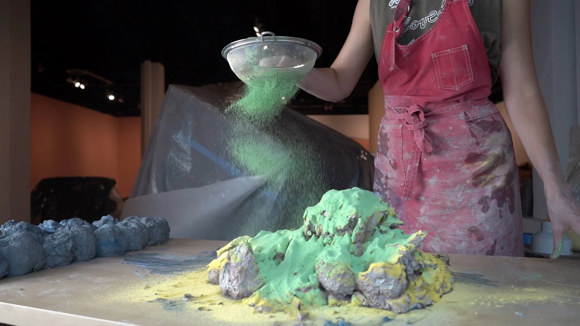 A person in a red apron is pouring green powder over a pile of mixed colored clay materials on a table in an art gallery setting.