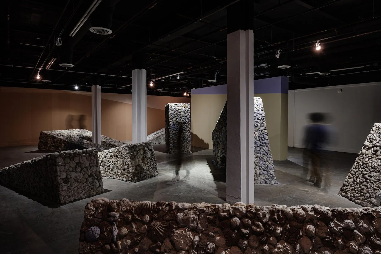 Contemporary art installation titled 'Future Pacific' featuring monumental sunken arches covered in clay forms like seashells and corals, displayed inside a gallery with dark ceiling, spotlights, and painted walls in orange, blue, and white.