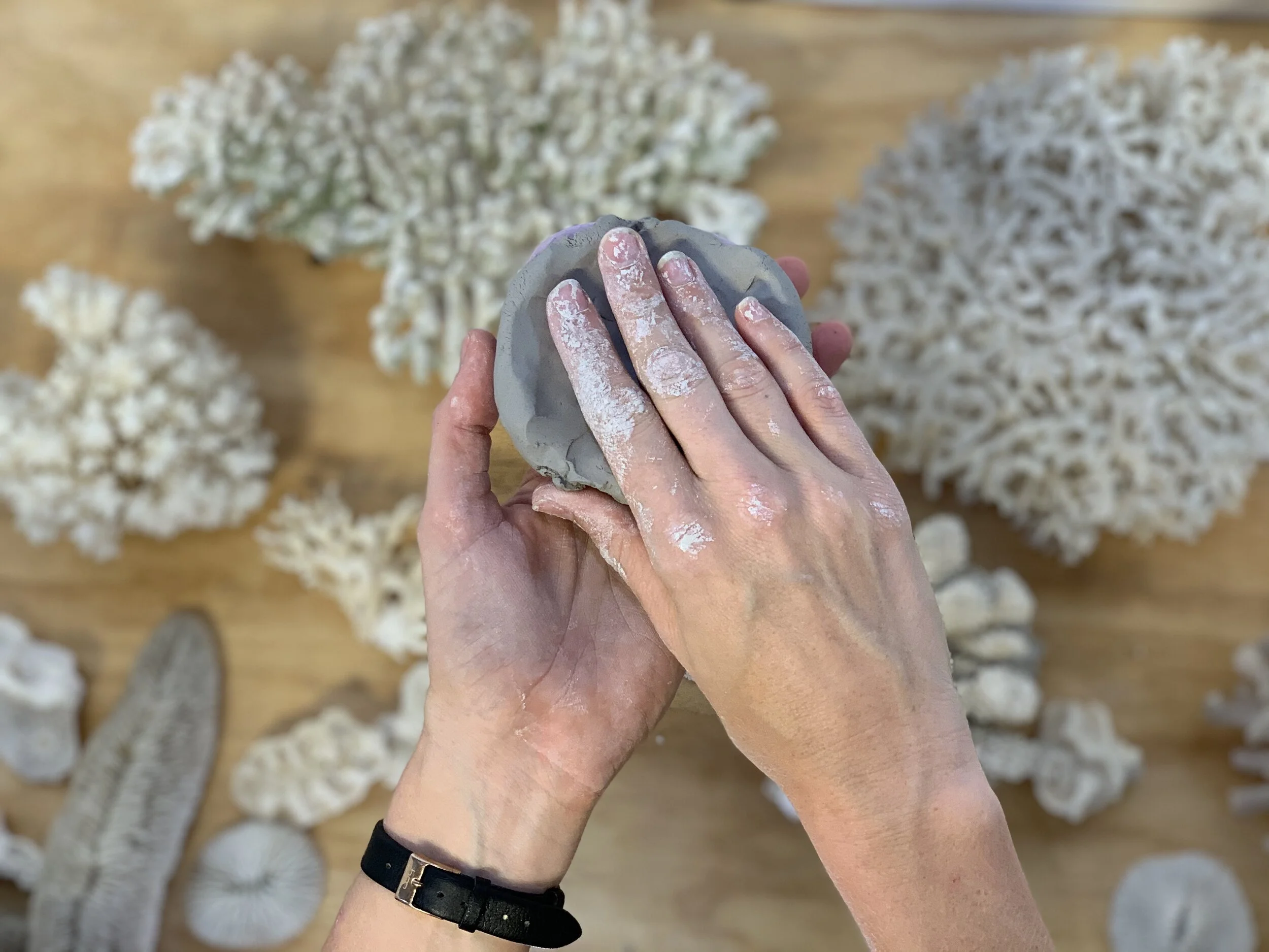 Person shaping a clay piece with both hands, surrounded by white coral pieces on a wooden surface.
