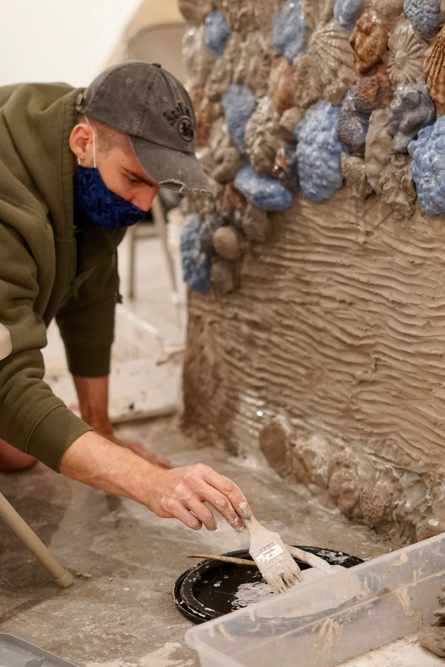 A person kneeling and painting a clay wall with a brush, wearing a gray cap, green hoodie, and blue face mask. The wall is covered in clay forms like seashells and corals.