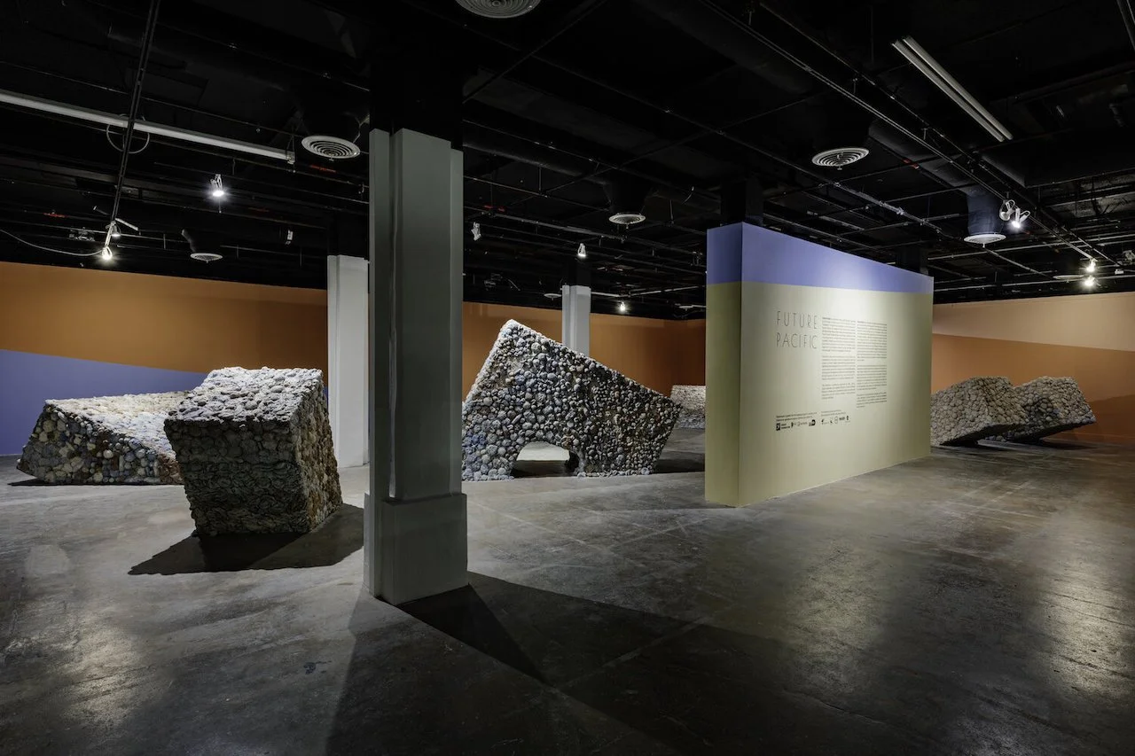 Contemporary art installation titled 'Future Pacific' featuring monumental sunken arches covered in clay forms like seashells and corals, displayed inside a gallery with dark ceiling, spotlights, and painted walls in orange, blue, and white.