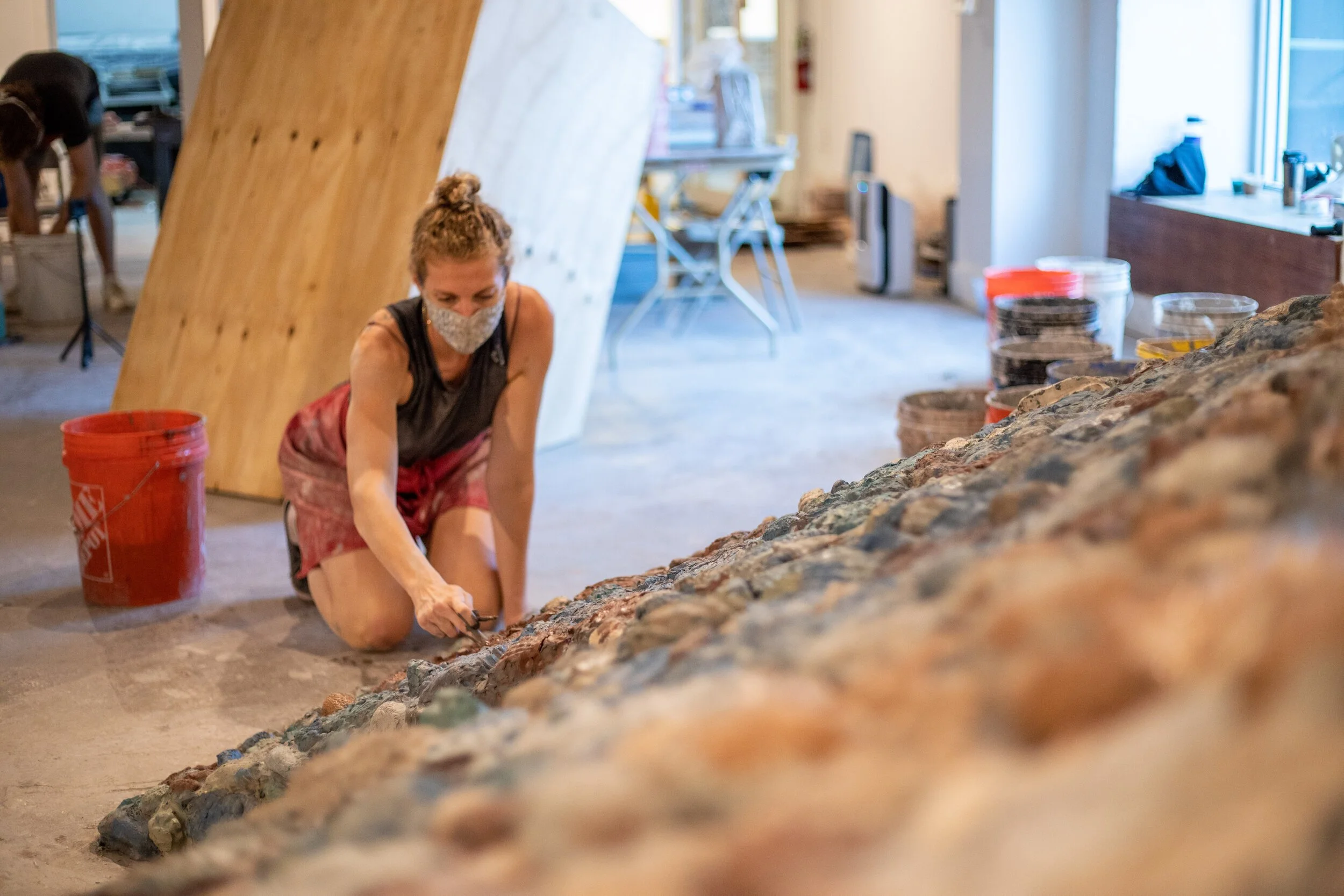 A woman kneeling and working on a clay wall in an indoor art gallery, wearing a mask, with buckets and wooden boards around.