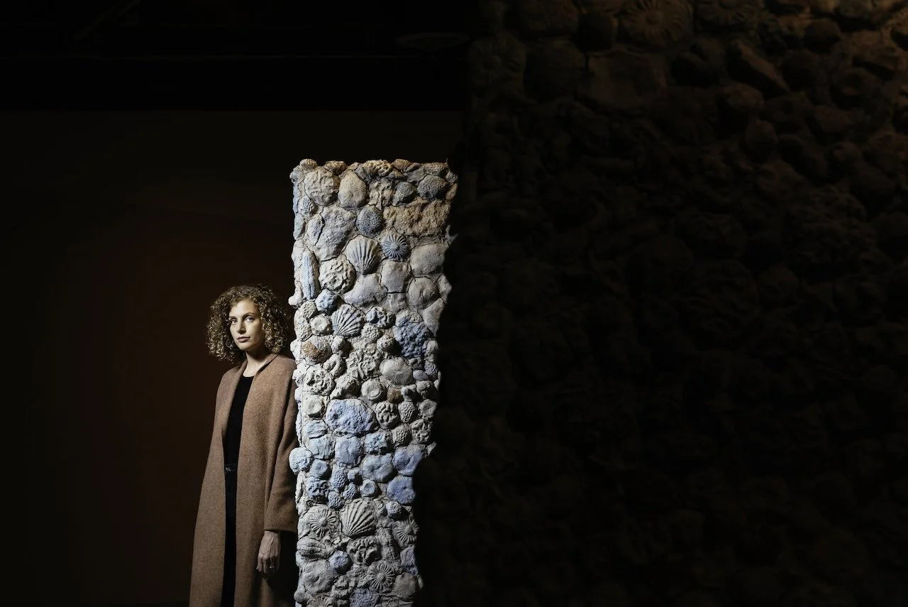 A woman with curly hair wearing a long beige coat standing beside a textured clay wall with coral and shell textures embedded in it, in a dimly lit setting.