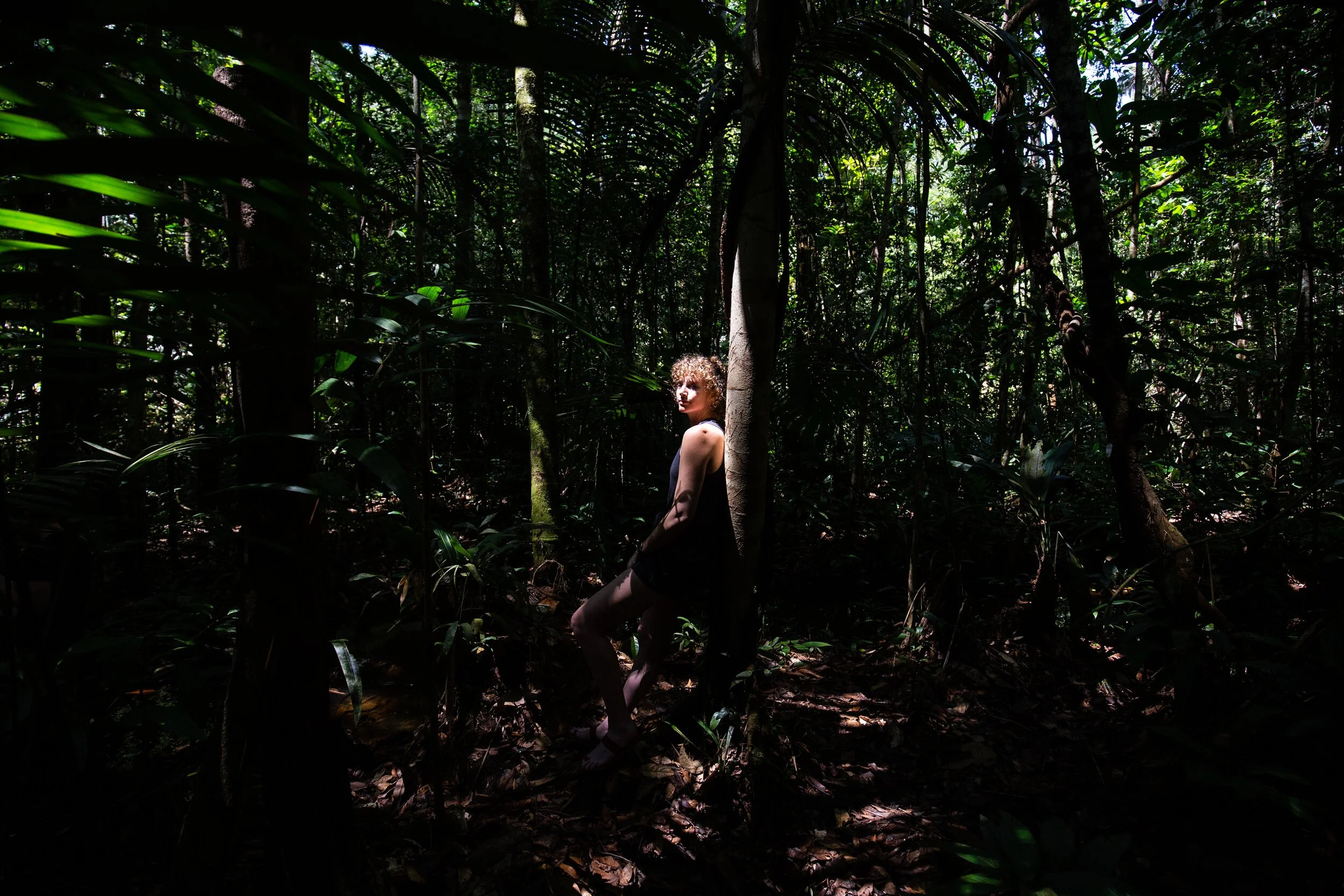 The artist, a woman with curly hair, pictured in a dark, dense rainforest with sunlight filtering through the canopy, leaning against a tree surrounded by tropical foliage.