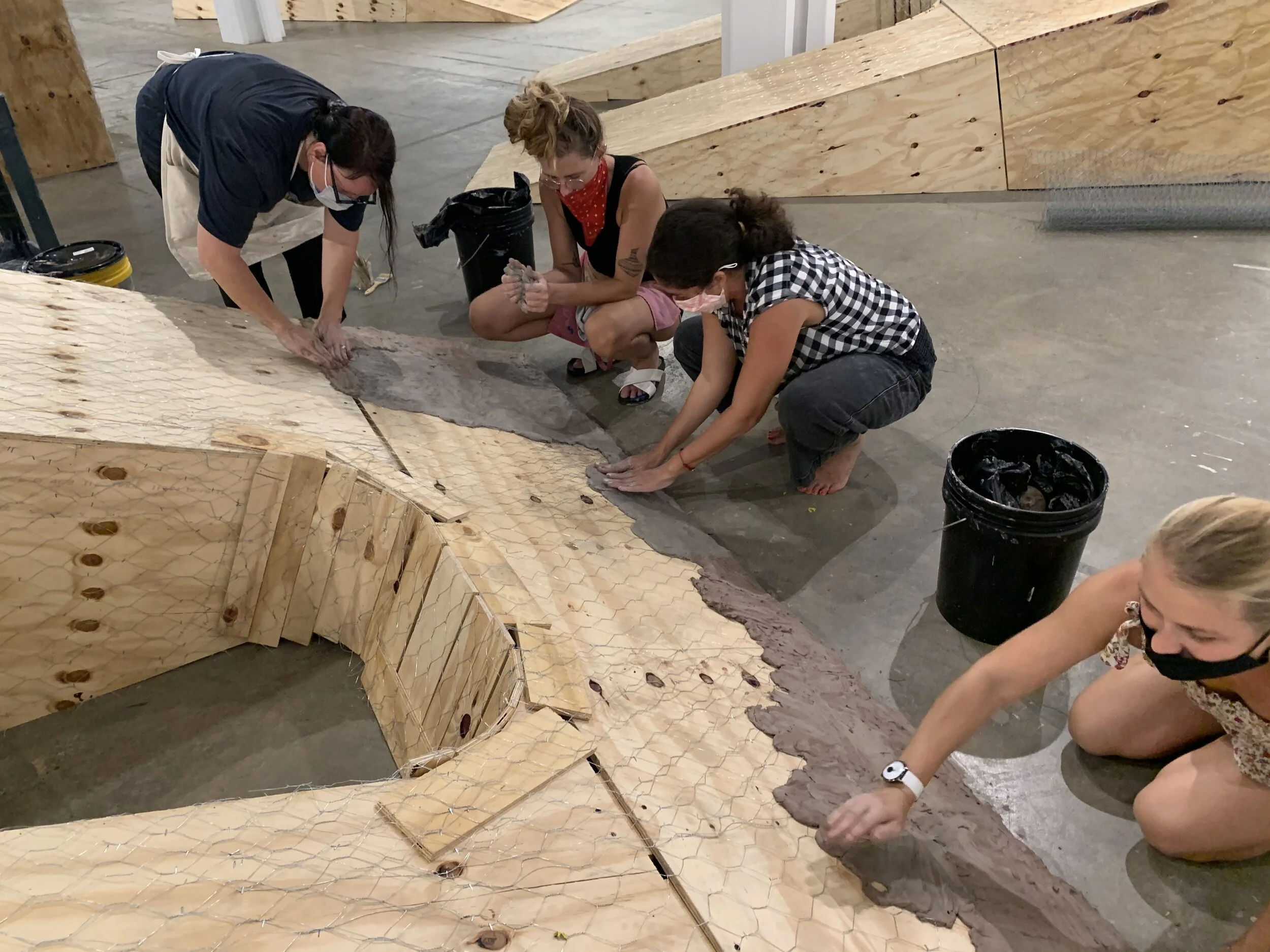Four women working together on a large wooden structure, applying a layer of clay on its surface inside the Bakehouse Art Complex art gallery.