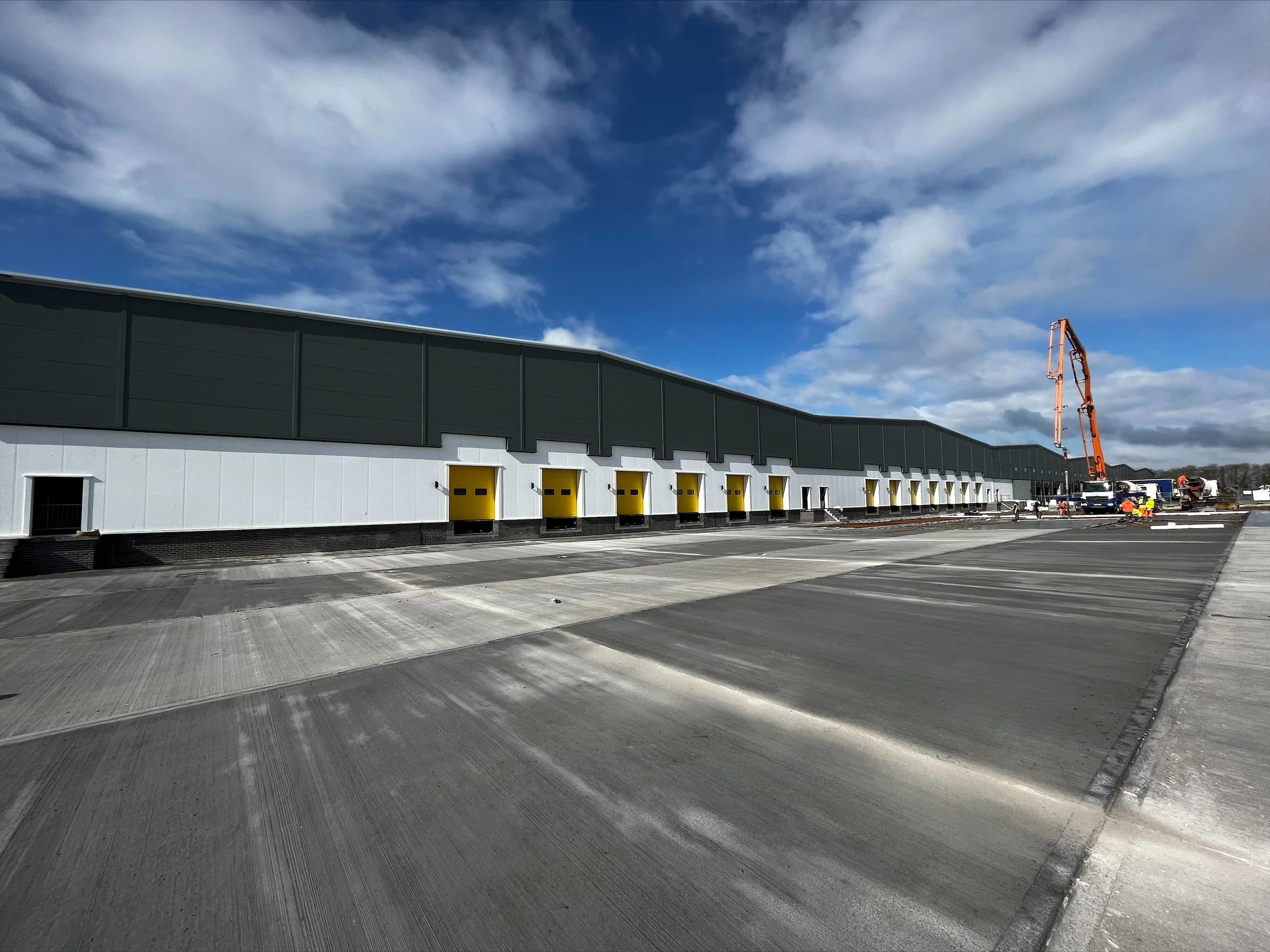 Large industrial warehouse with yellow loading dock doors, concrete loading bay, and construction equipment under a cloudy sky.