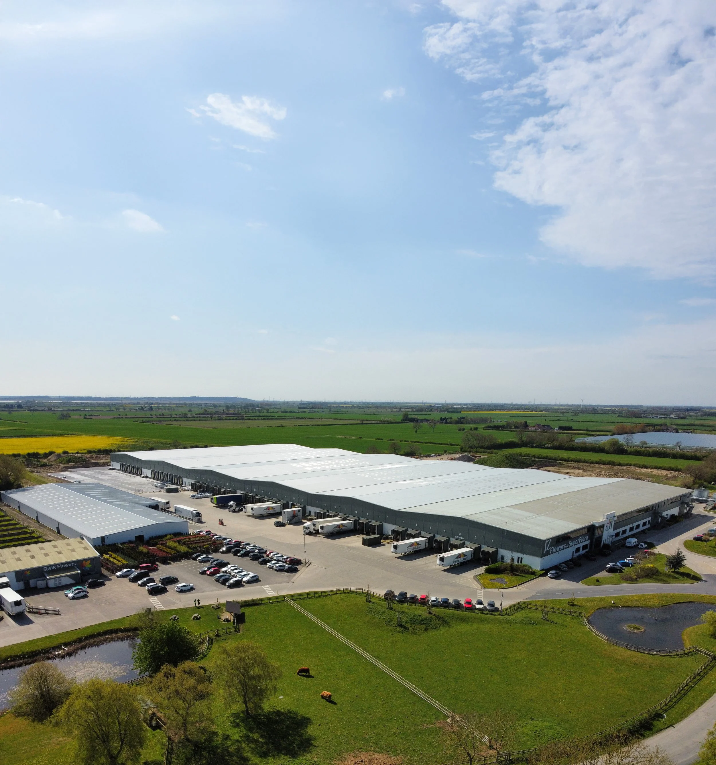 Aerial view of a large warehouse complex with delivery trucks surrounded by green fields and farmland under a partly cloudy sky.