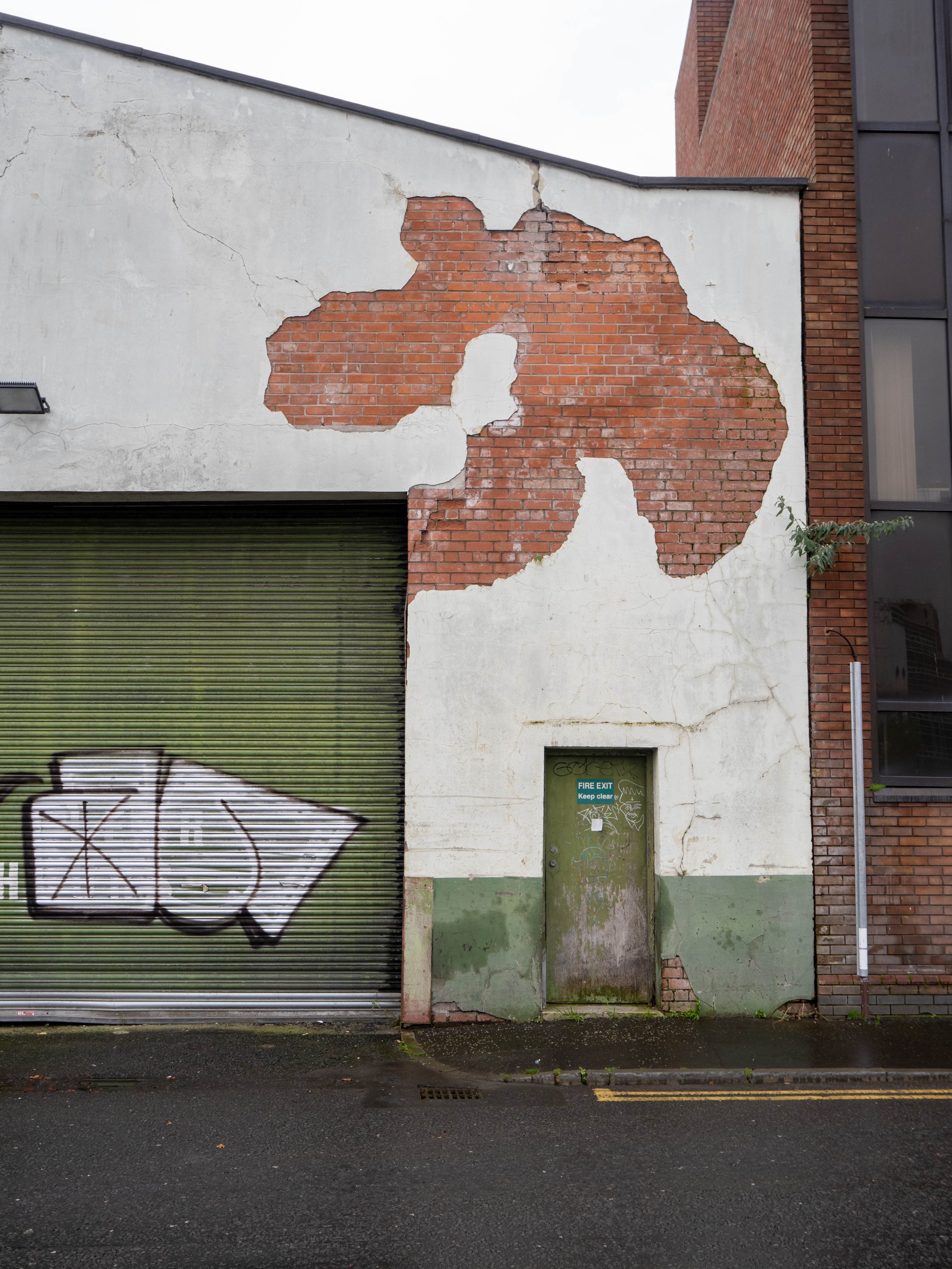 A weathered building wall with peeling white paint revealing red brick underneath, alongside a green garage door with graffiti, and a green door with a 'Fire Exit' sign.