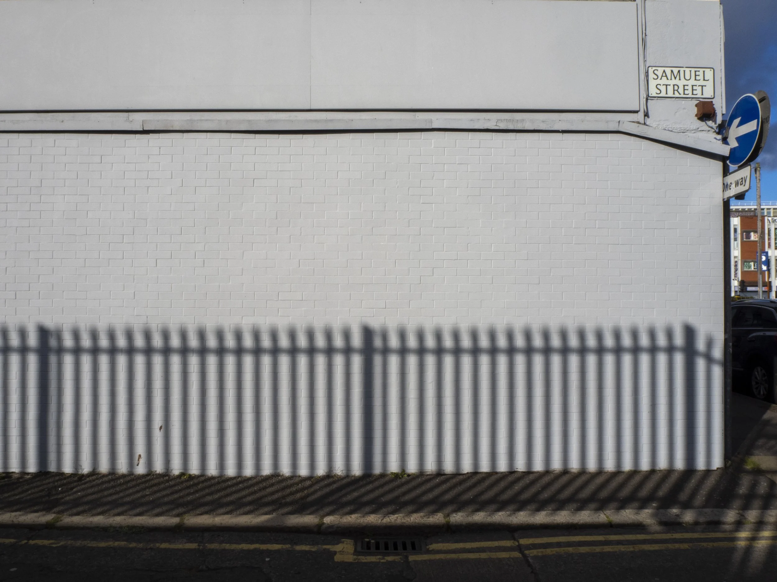 A white brick wall with a shadow of a metal fence cast on it, a street sign reading 'Samuel Street' and a one-way street sign on the corner, with a blue sky and part of a cityscape in the background.