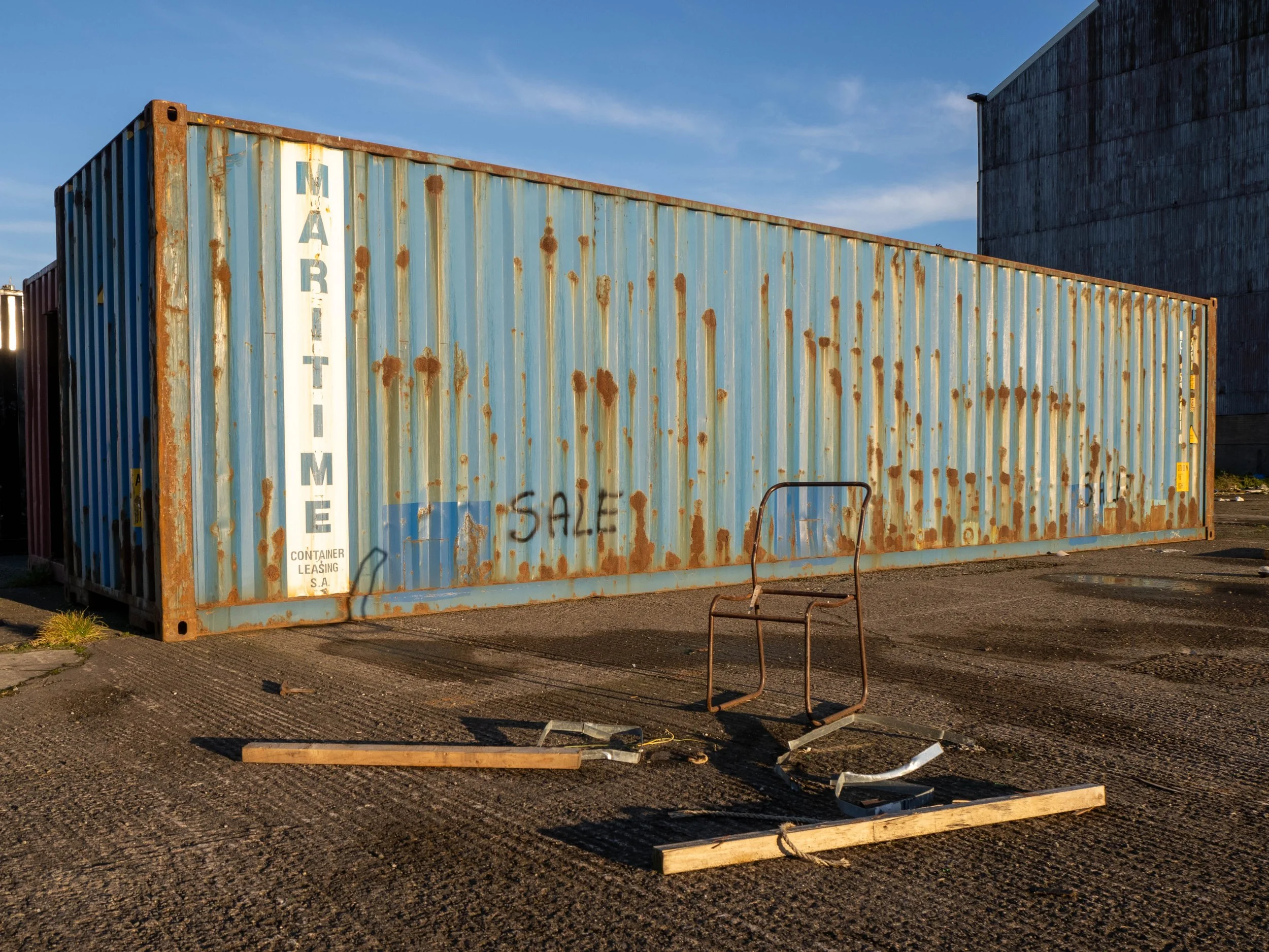 Old, rusty blue shipping container with graffiti sale sign on the side, on a gravel lot with scattered broken wood and metal debris, and a weathered building in the background.