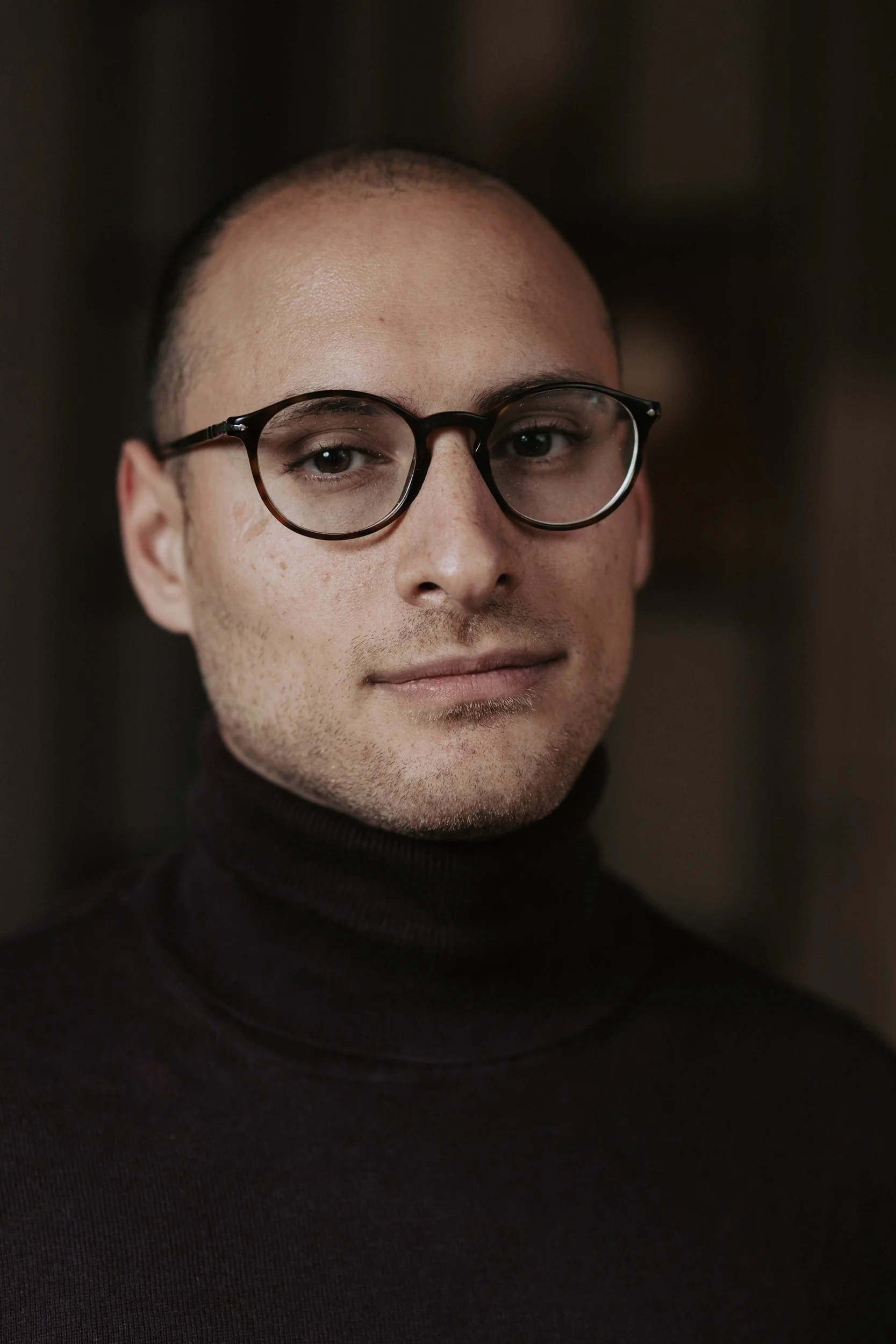 Ritratto di un uomo con occhiali e maglione a collo alto, sfondo sfocato.