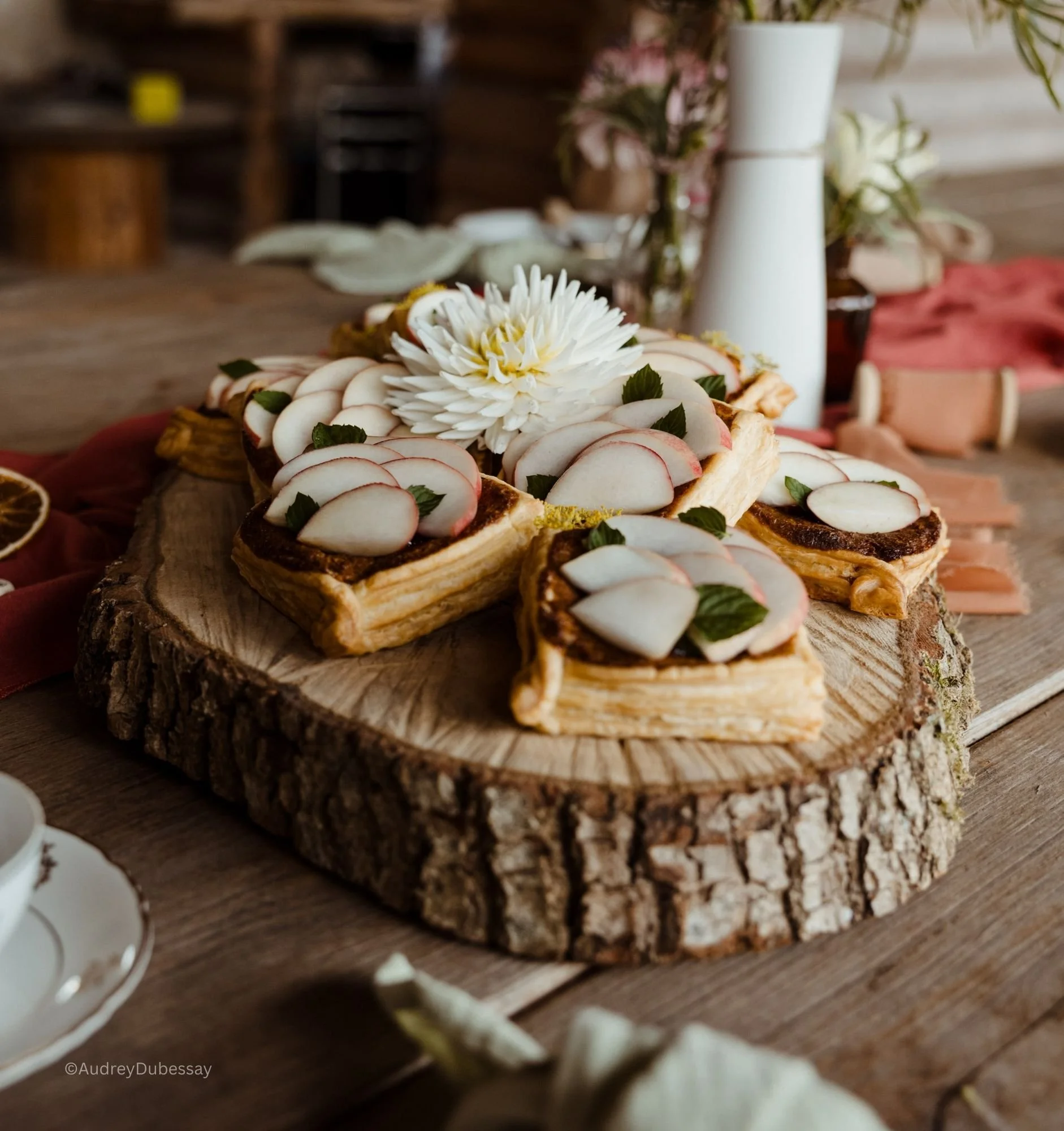Gâteaux à la crème avec des tranches de pomme, décorés de feuilles de menthe, disposés sur une tranche de bois avec une grande fleur blanche en décoration.