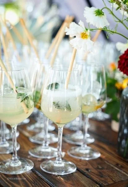 Verres à cocktail avec garnitures de menthe et citrons, décorés de bâtons de bois, et fleurs blanches et rouges en arrière-plan sur une table en bois.