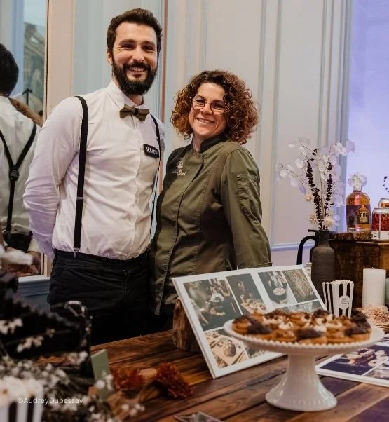 Deux personnes souriantes, un homme et une femme, posent à côté d'un gâteau sur une table dans une pièce élégante, avec des livres et des décorations sur la table.