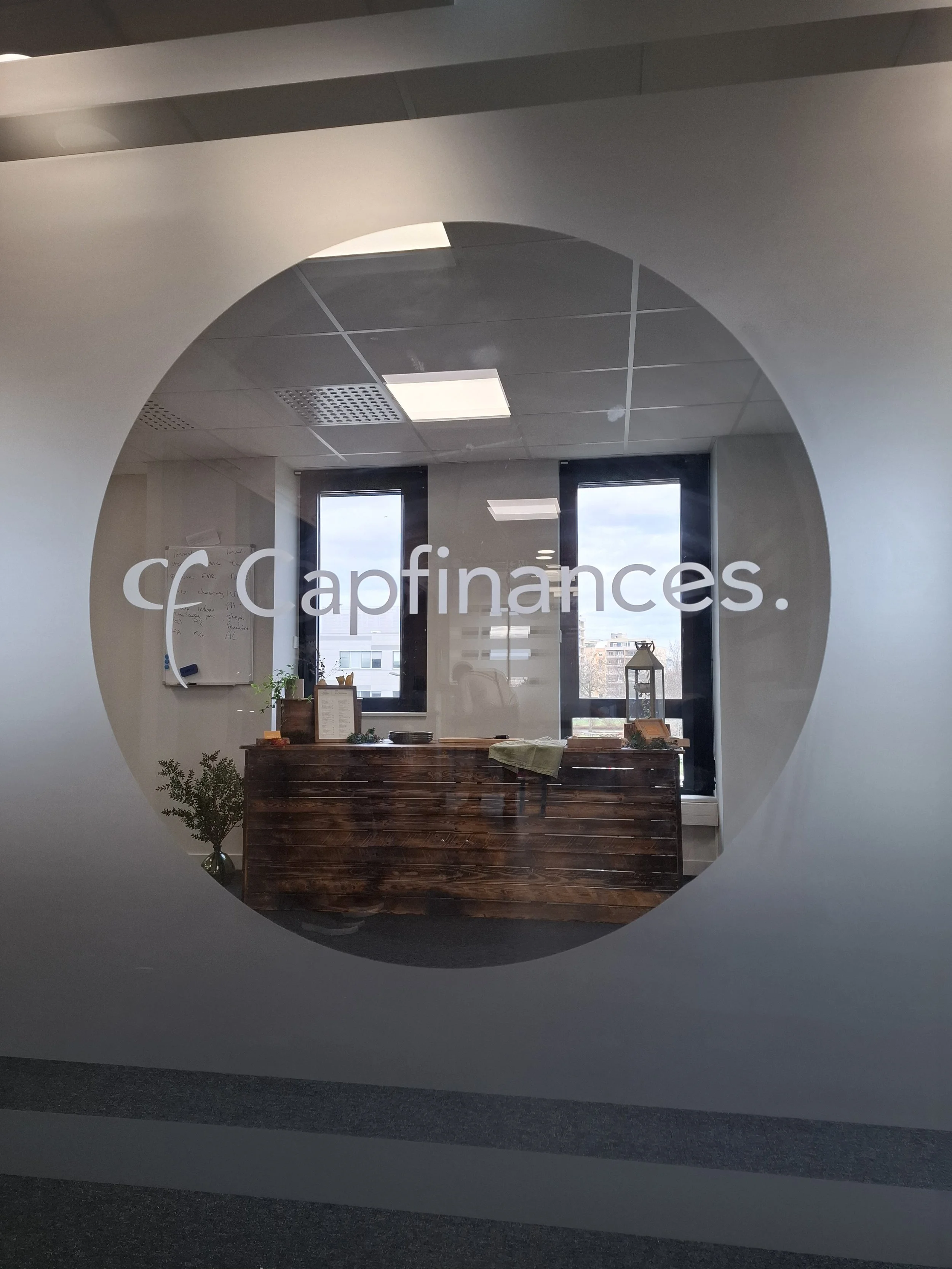 Vue à travers une fenêtre avec un cercle découpé, affichant un bureau avec une réception en bois foncé, une plante, et une décoration d'intérieur, avec deux grandes fenêtres en arrière-plan.