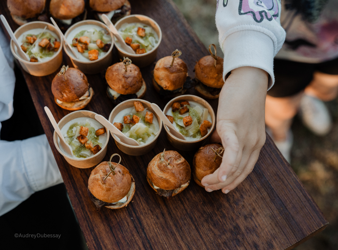Petits sandwiches avec bun brioché, servis avec des cuillères en bois contenant une crème verte garnie de croûtons. La personne manipulate les sandwiches.