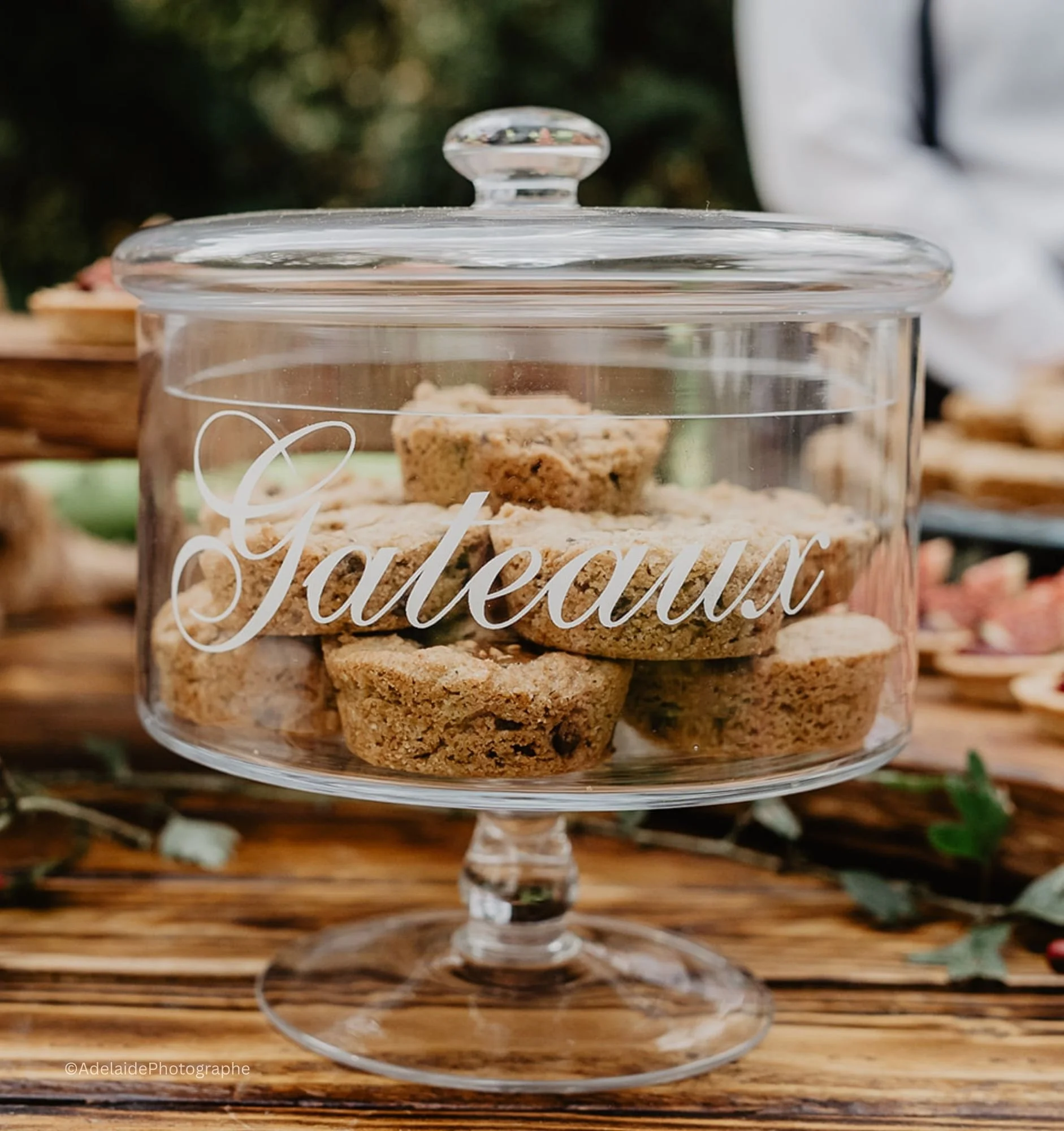 Un contenant en verre avec des biscuits étiqueté 'Gâteaux' placé sur une table en bois, avec d'autres pâtisseries en arrière-plan.