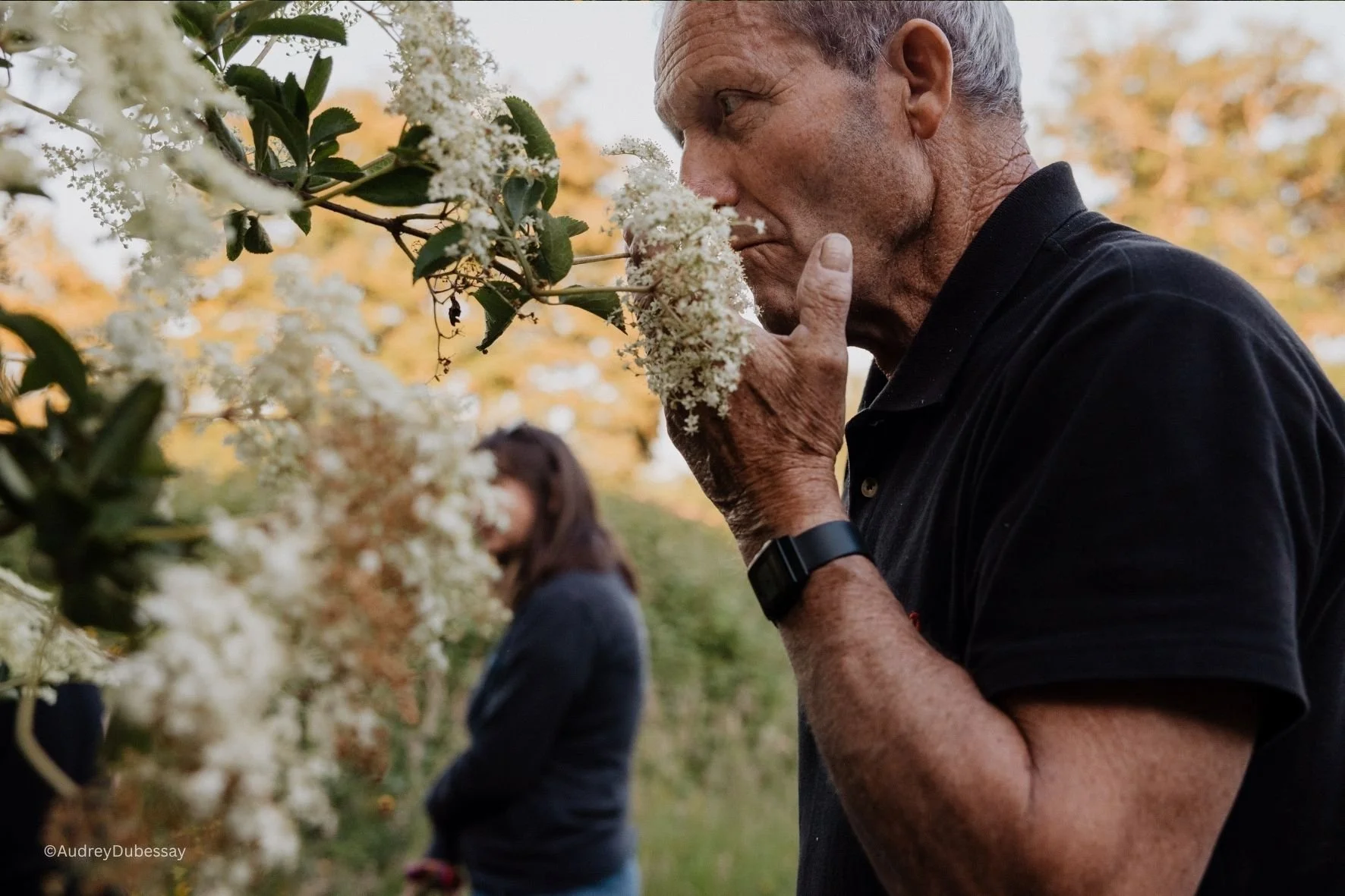 Un homme âgé sentant une fleur blanche dans un jardin avec des arbres aux feuilles orange en arrière-plan, accompagné d'une femme en arrière-plan.