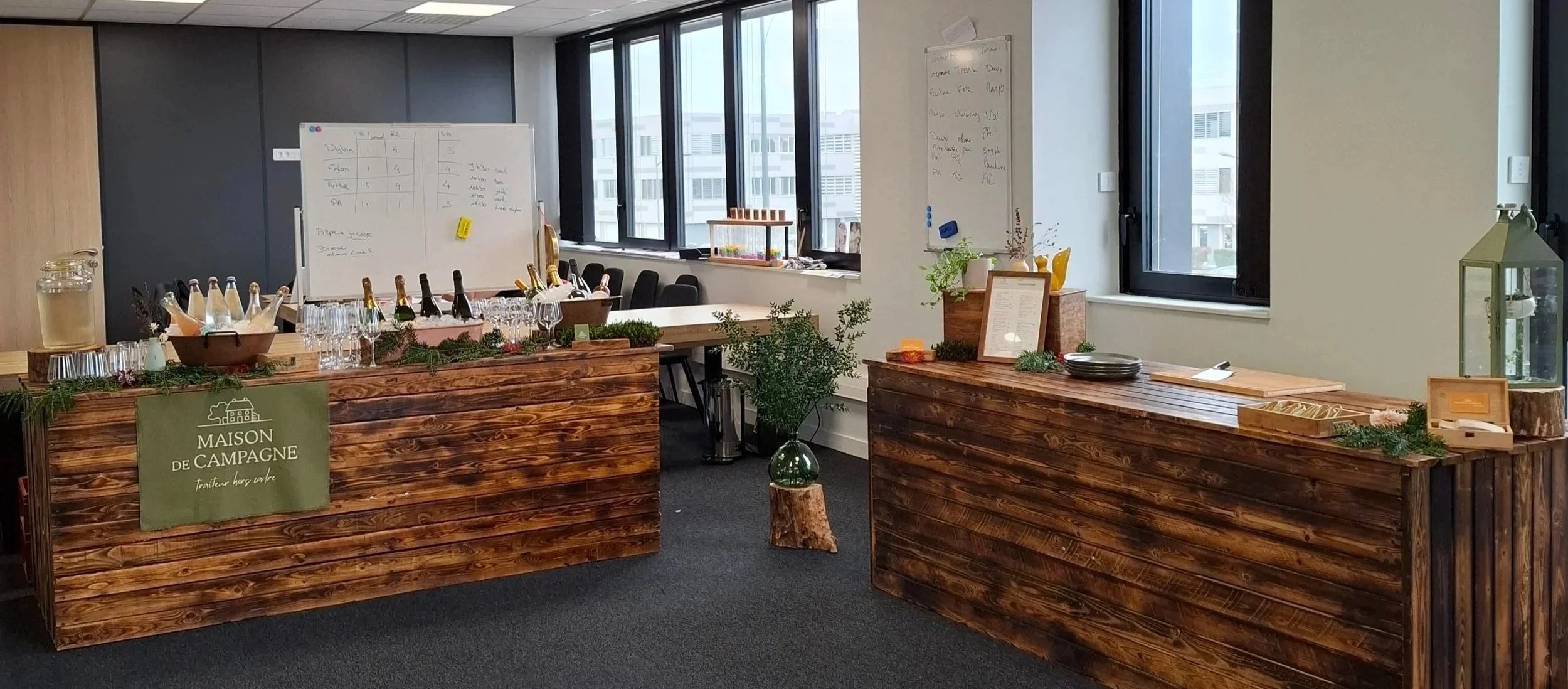 Table rustique en bois avec quelques décorations et vaisselle, espace de réunion ou de dégustation dans une salle moderne avec grandes fenêtres, décorée de feuillage et d'éléments naturels.
