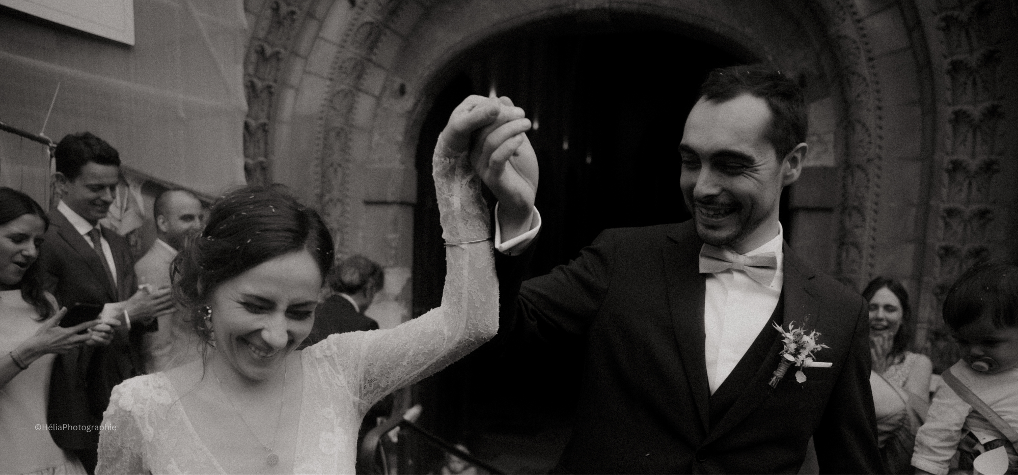 Un couple marié danse lors de leur réception dans une église ancienne, entourés de proches. La mariée porte une robe en dentelle et le marié un costume noir avec une lavallière. La scène est en noir et blanc.