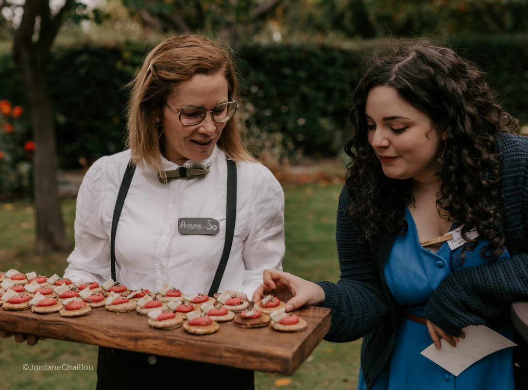 Une serveuse souriante, portant un uniforme blanc avec un badge nommé 'Ariane So', présente un plateau avec des biscuits décorés, tandis qu'une cliente en bleu regarde et touche un biscuit lors d'un événement en plein air.