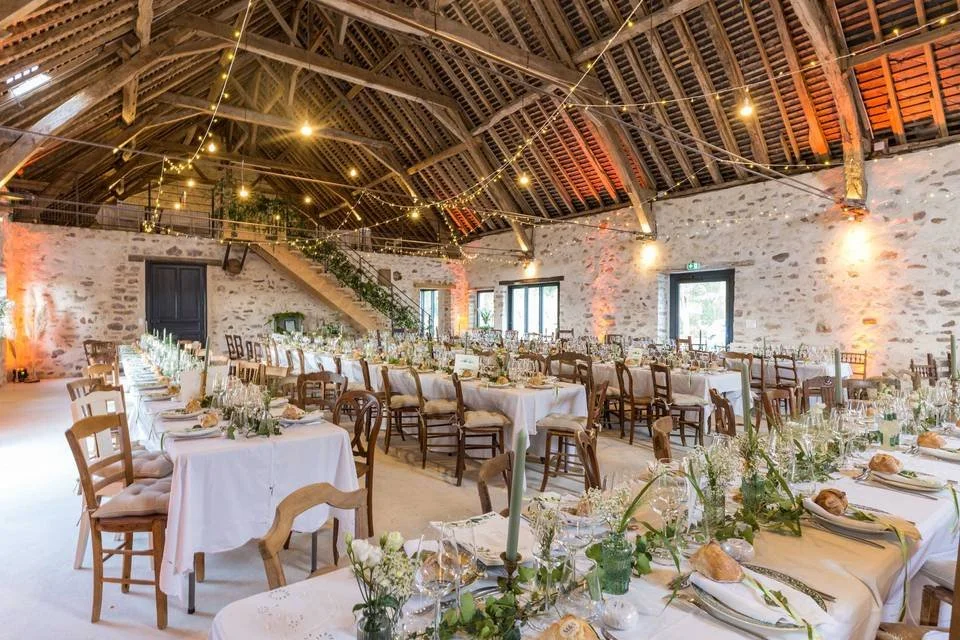 Salle de réception décorée pour un mariage ou un événement, avec des tables aménagées, guirlandes lumineuses suspendues, murs en pierre et plafond en bois.