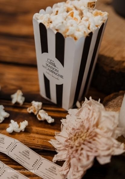 Un bol de popcorn dans un récipient rayé noir et blanc, posé sur une table en bois, avec une fleur pâle et un ticket de cinéma à côté.
