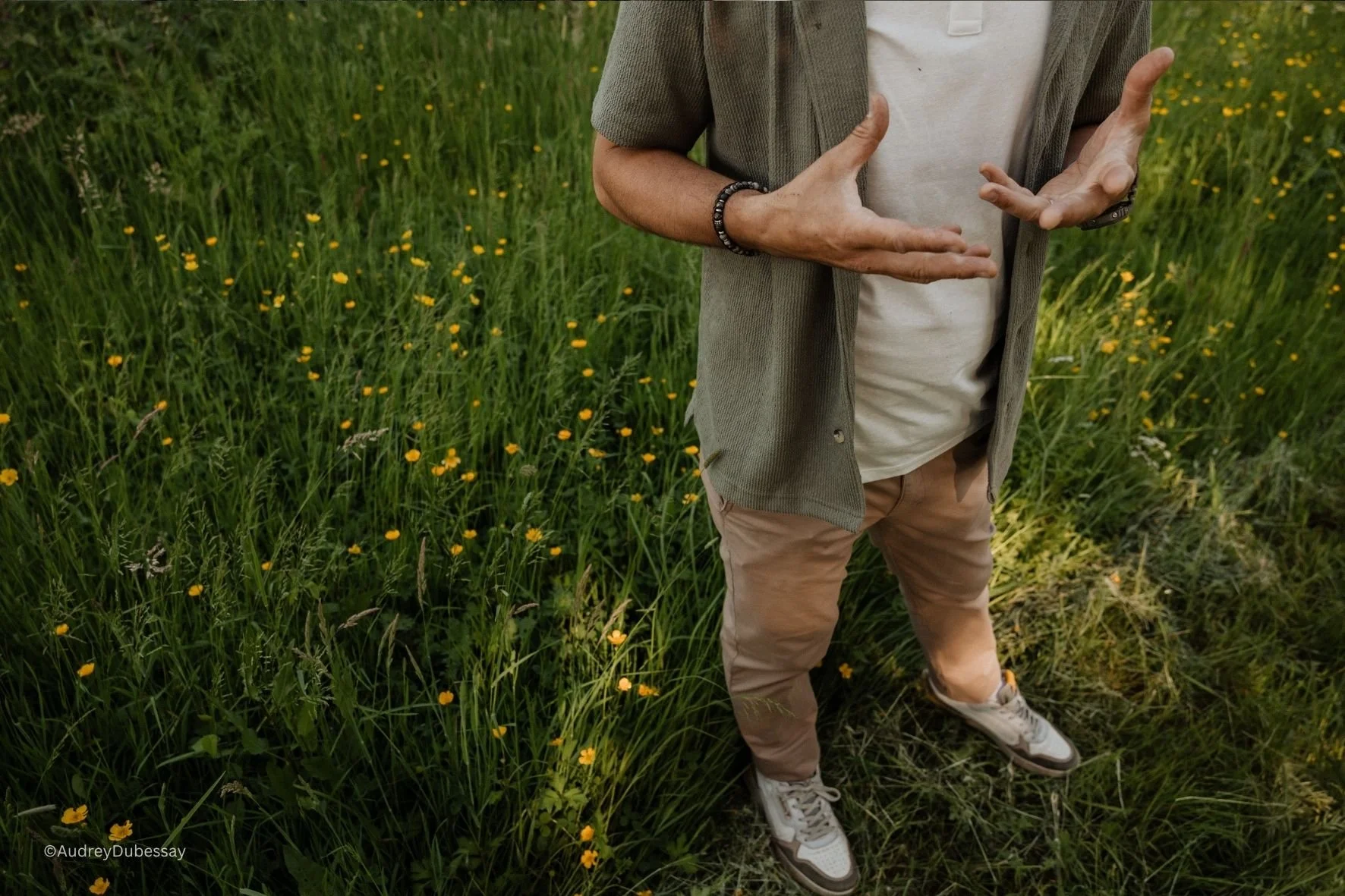 Personne debout dans un champ d'herbes vertes avec de petites fleurs jaunes, portant un t-shirt blanc, un pull gris léger, un jean beige et des chaussures blanches, gesticulant avec les mains.