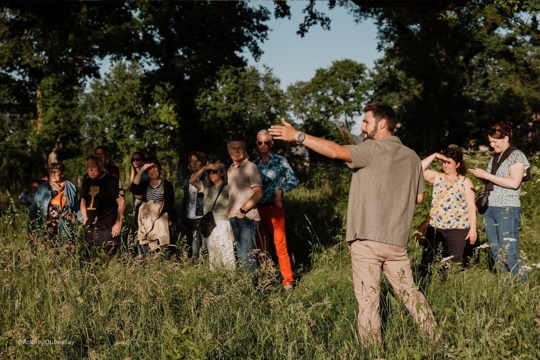 Un groupe de personnes assises ou debout dans un espace extérieur herbeux, écoutant un guide ou un conférencier parlant avec un geste de la main, sous un ciel clair avec des arbres en arrière-plan.