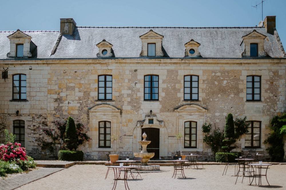 Un grand bâtiment en pierre avec plusieurs fenêtres et un toit en pente recouvert de tuiles, un fontaine centrale dans une cour avec des tables et des chaises, des plantes et des fleurs